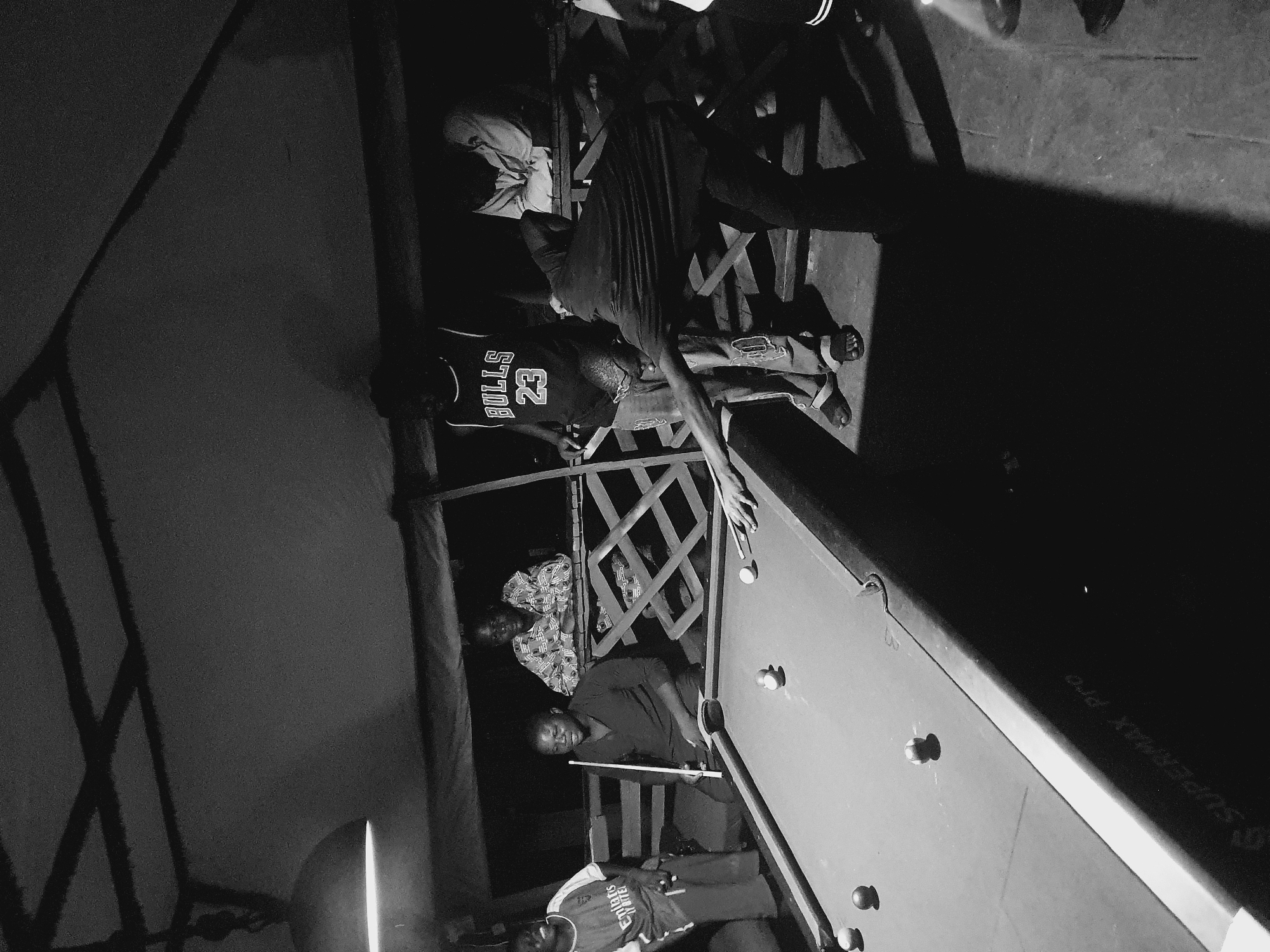In a dimly lit interior, potentially in Lokoja, Nigeria, several individuals engage in a game of pool on a "SUPERMAX PRO" branded table. One man, wearing a "BULLS 23" jersey, stands observing the game. Another man, dressed in a "CHELSEA" jersey, is actively playing. A group of people are also observed standing on a balcony watching the game, constructed with wooden railings and beams, positioned behind the players.