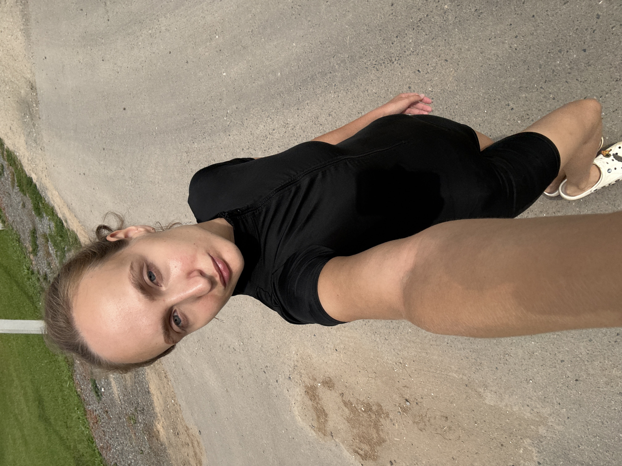 The media file shows a person lying down on a paved surface, possibly a road or pathway. The person is wearing a black t-shirt and shorts, and white clogs with a cartoon design. Their right arm is extended towards the camera, creating a selfie-like perspective. The person has fair skin and light-colored hair, and their eyes are open and looking at the camera. Their facial expression appears neutral.
The setting is outdoors, with a gray paved ground and a patch of green grass and some gravel visible in the background. The lighting suggests it is daytime, possibly late afternoon or early evening, given the quality of light. There is no visible text in the image. The overall impression is of a casual moment captured outdoors. The location context provided is Подшивалово, Russia, which helps to identify the geographical setting of the image.