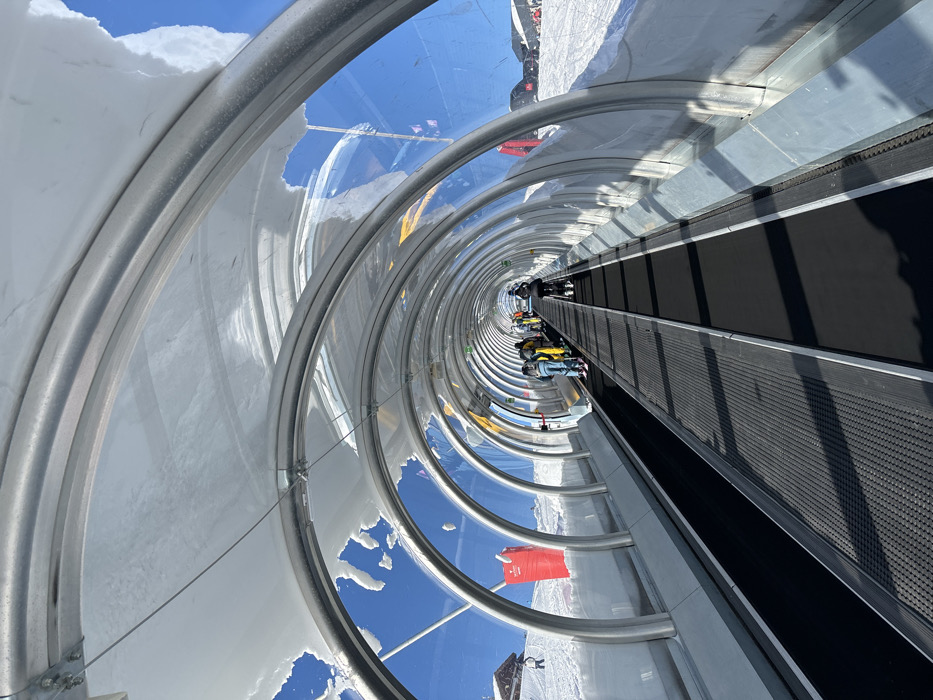 This vibrant image depicts the interior of a transparent, enclosed conveyor belt ski lift, often referred to as a "magic carpet," at a ski resort in Val-d'Isère, France. The perspective looks down the long, tubular structure, which is framed by repeating silver-colored metal arches and clear panels, creating a strong sense of depth.
Several individuals in winter ski attire, including one person in a prominent yellow jacket, are visible on the black conveyor belt, being transported uphill. Some are on skis, while others are standing. To the right of the moving belt, a stationary, textured black surface serves as a pathway or queuing area.
The transparent walls offer panoramic views of the bright blue sky and the surrounding snow-covered mountain landscape. Patches of snow cling to the exterior and interior edges of the transparent panels, particularly on the left side, indicating the alpine environment. The scene is brightly lit by natural sunlight, suggesting a clear, sunny day, with distinct shadows cast on the conveyor belt and the snow outside the tunnel. A red sign with illegible white text is partially visible outside the tunnel structure, likely indicating a safety message or information. The image captures modern ski infrastructure designed for convenient and scenic ascent in a snowy mountain setting.