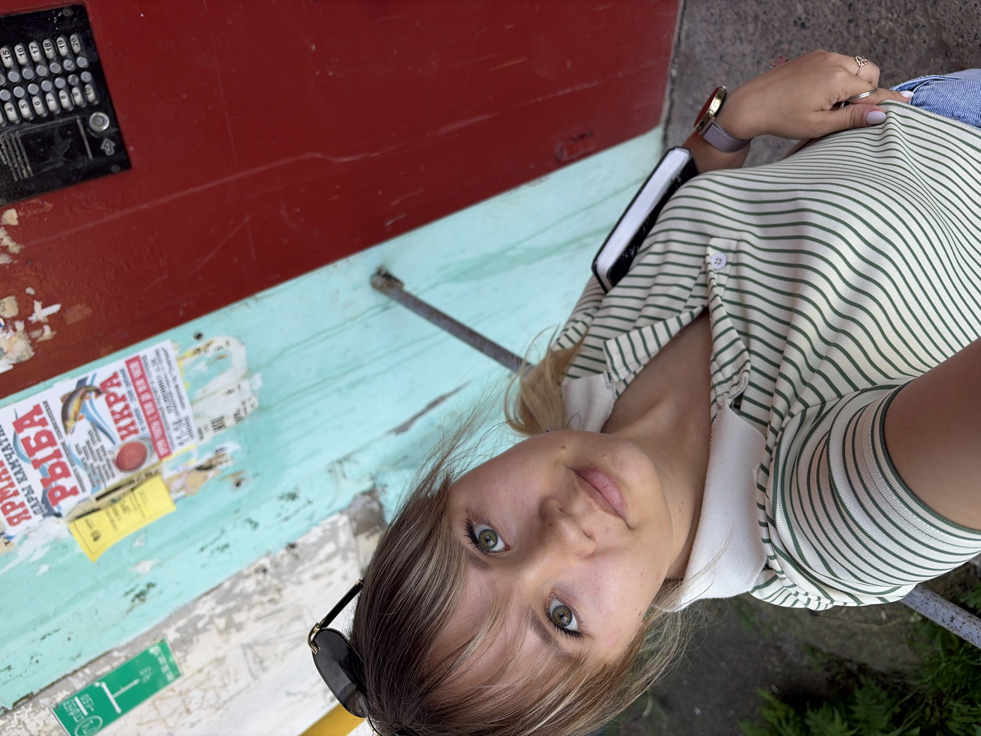 In Chaikovsky, Russia, a young woman with blonde hair and green eyes, wearing a striped polo shirt and sunglasses, is captured in a close-up, selfie-style photograph. She is positioned in front of a weathered, light blue wall adorned with various posters. To her right, a bright red door with a black intercom panel is visible. The ground is a rough, concrete surface. The woman's left arm is slightly visible, and she is wearing a watch on her left wrist and rings on her fingers. A dark object, possibly a book or tablet, is tucked into her shirt. The lighting suggests an outdoor setting during daylight hours.