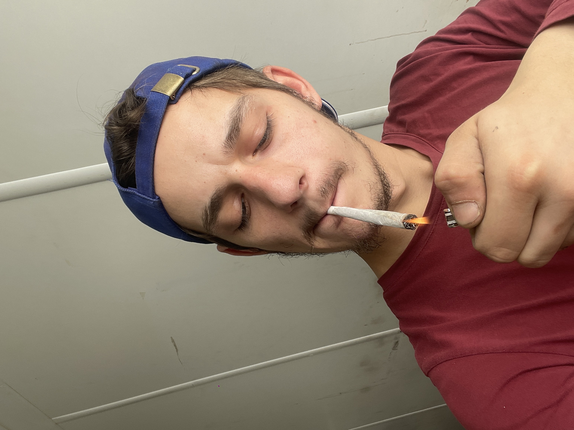 A person wearing a red t-shirt and a blue cap is lighting a rolled cigarette with a lighter. The location appears to be indoors with a white wall and pipe visible in the background.