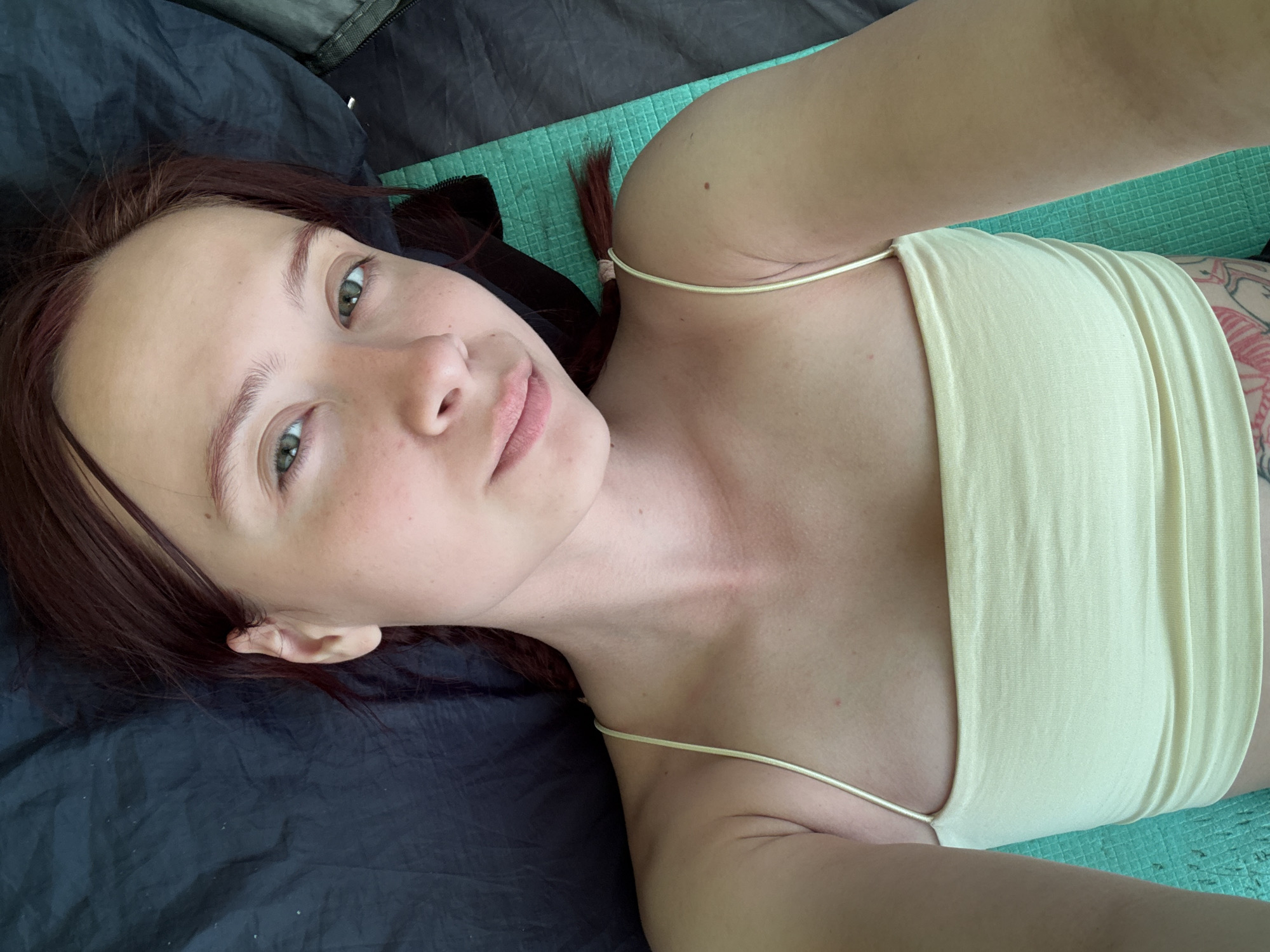 A selfie taken from an overhead angle shows a person lying down. The person is fair-skinned with reddish-brown hair styled in pigtails. The person wears a light yellow top with thin straps. Tattoos are visible on the right side of the torso. The person is lying on a dark gray surface to the left and a light green surface to the right.
The location is unknown.