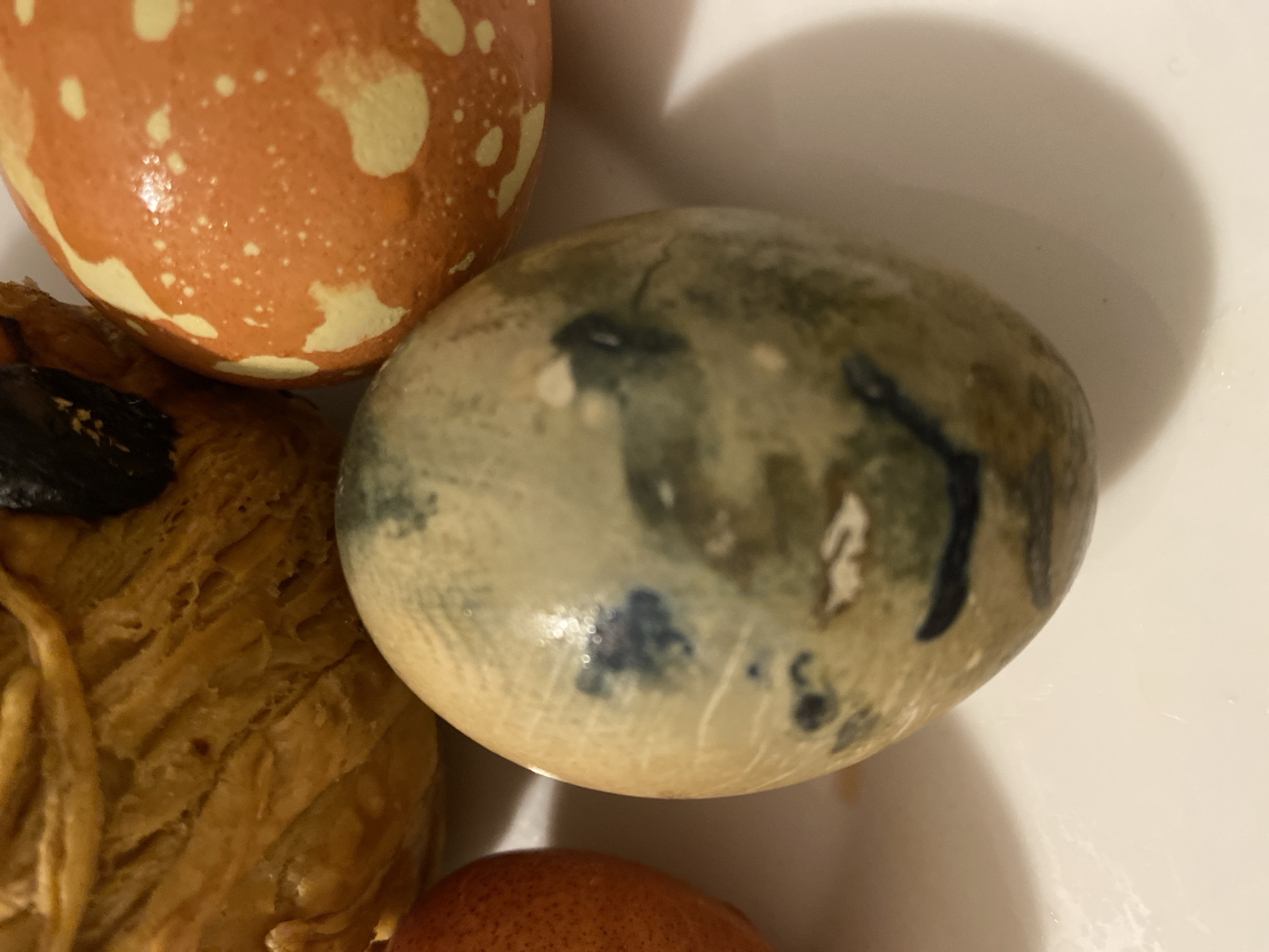 The image shows a close-up of three painted Easter eggs and a piece of pastry on a white surface. One egg is orange with white spots, another is light brown with dark blue and green marbling, and a third smaller egg is reddish-orange. The pastry appears golden brown and textured. The setting does not provide information to determine if this image was taken in Alanya, Turkey, or any other location.