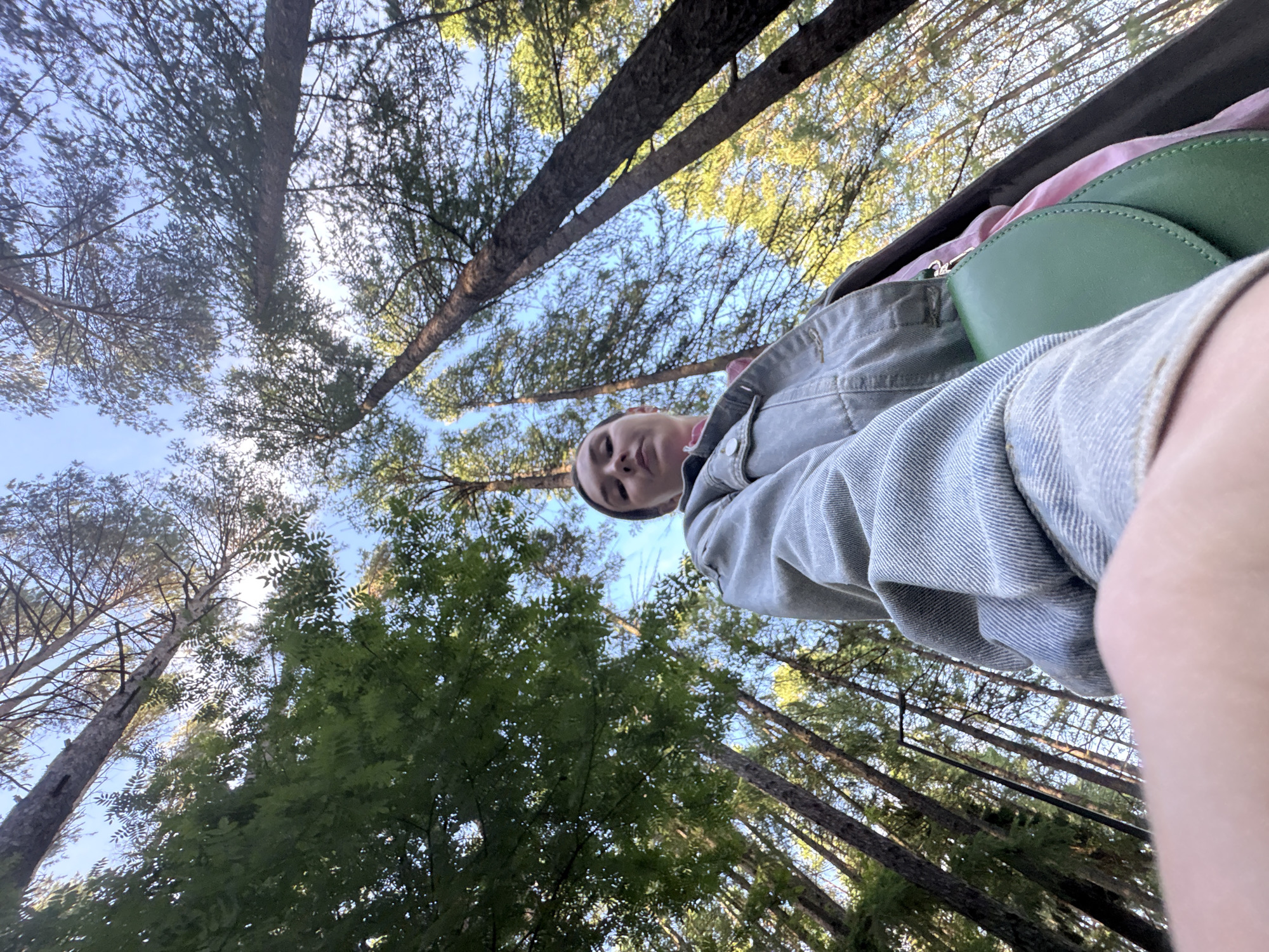 The media file is a low-angle, wide-shot selfie taken from a person lying down in a forest. The main subject is a person, partially visible from the chest up, wearing a denim jacket and a pink top. They are looking upwards towards the camera with a neutral expression. A green purse is visible on top of their chest.
The setting is a forest with tall trees, primarily pines, with some deciduous trees also present, judging by the lush green foliage. The camera is pointed directly upwards, capturing the canopy of the trees against a clear blue sky. The sunlight filters through the branches, creating a dappled light effect. The time of day appears to be late afternoon or early evening, given the angle of the sun and the warm tones of the light. The weather seems pleasant and clear.
The person appears to be relaxing or resting in the forest. The low angle of the shot emphasizes the height of the trees and the open sky, creating a sense of being immersed in nature. There is no visible text in the image. The location is identified as Izhevsk, Russia, suggesting an outdoor recreational activity in that region.