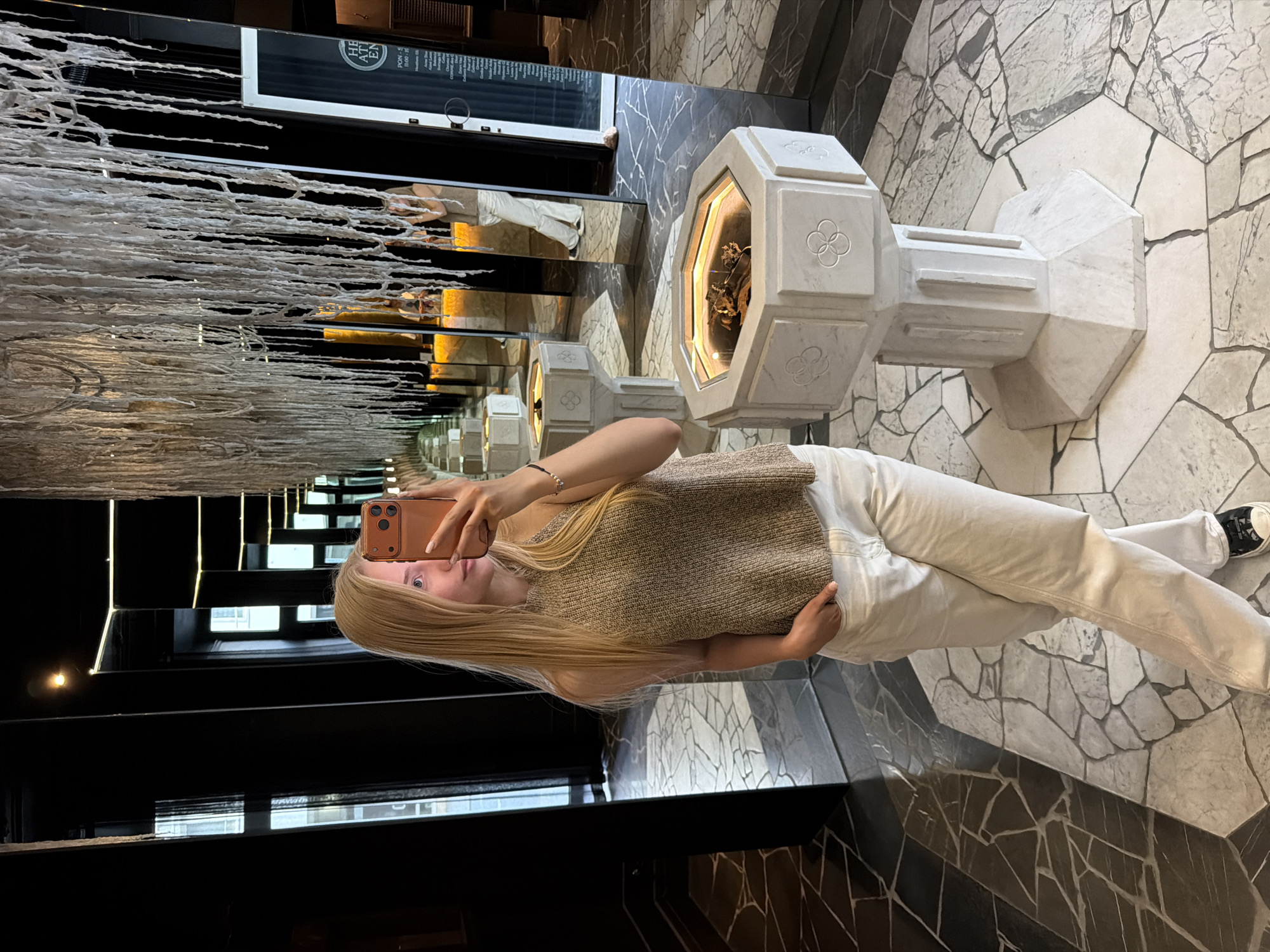 The image displays a young adult female taking a selfie in an indoor setting with extensive use of mirrors, creating an illusion of infinite depth. The location is Unknown, Unknown. The female has long, straight blonde hair and is wearing a light brown, possibly knit, sleeveless top, light-colored (likely off-white or cream) pants, and black and white low-top sneakers. She holds an orange-colored smartphone with a visible triple-camera array in her right hand, aiming it towards herself, while her left hand is casually placed in her pants pocket. A delicate bracelet is visible on her left wrist.
The environment features dark, reflective walls, predominantly covered by tall mirrors that generate multiple reflections of the room, the subject, and other indistinct figures in the background. An elaborate ceiling installation composed of numerous white, string-like or textured organic forms hangs above. The floor is paved with an irregular mosaic of light and dark grey stone tiles. Throughout the space, and receding into the reflections, are multiple white, hexagonal stone pedestals, each featuring a central illuminated display containing what appears to be dried organic matter. These pedestals are adorned with quatrefoil emblems. A reflected sign in the background partially displays text including "HE AT EN" and "PON-S 11:00 - 18". Windows showing an external view are also visible within the mirrored reflections.