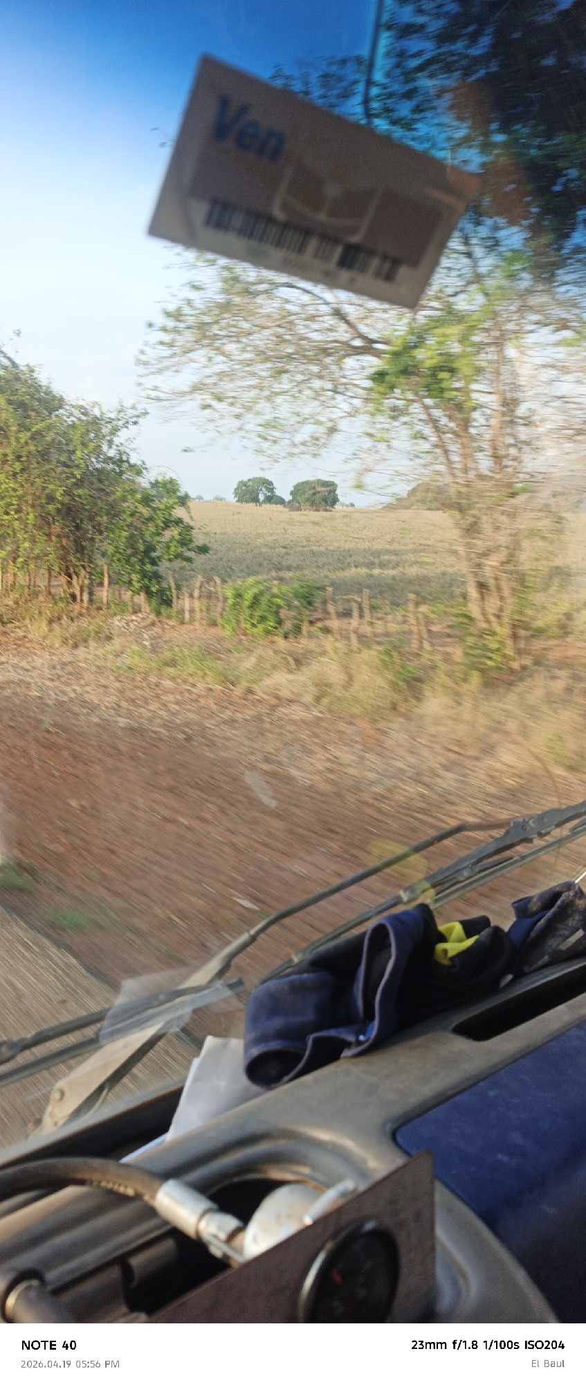 The image, taken from inside a moving vehicle, shows a rural landscape in Venezuela. In the foreground is the vehicle's dashboard, with a blue cloth and various car parts visible. Through the windshield, a countryside scene unfolds, featuring a field with sparse vegetation, a wooden fence, and scattered trees. A sticker with the word "Ven" is attached to the windshield, partially obstructing the view. The photograph appears to be taken in motion, causing some blurring of the scenery. According to the included text, the location is El Baul, Venezuela.
