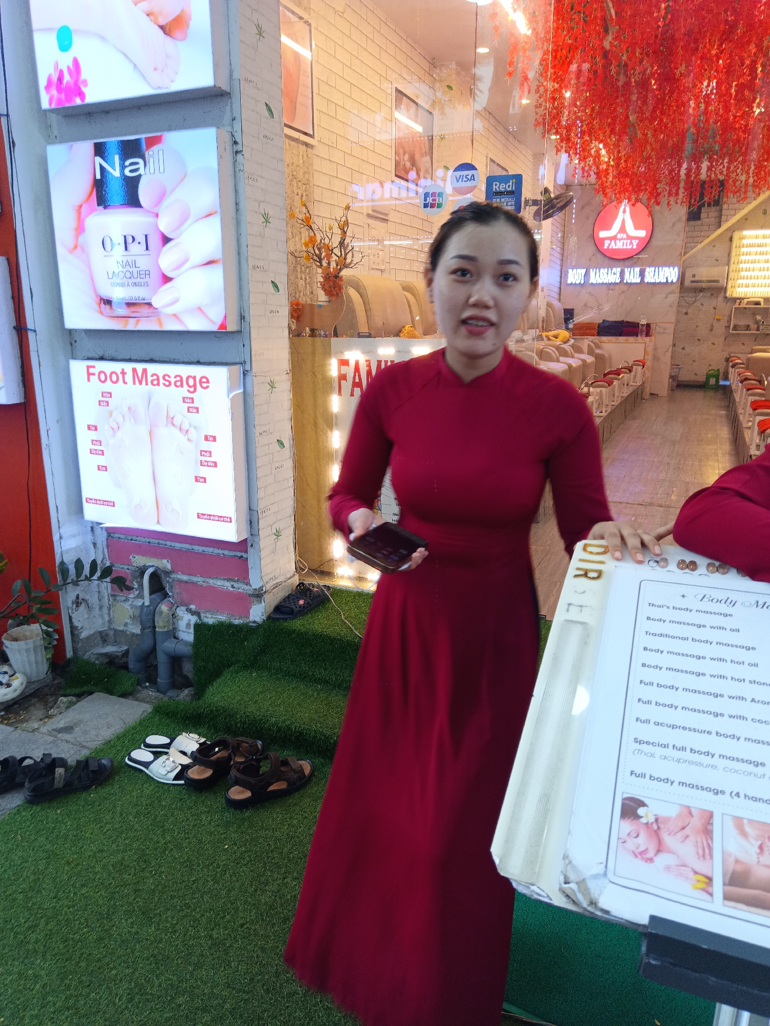 The photograph depicts a woman in a red dress standing outside a spa in Nha Trang Ward, Vietnam. She is holding a phone in one hand and resting the other on a sign displaying a menu of massage services. The sign lists various massage types, including Thai, aromatherapy, and coconut massages, along with a "special full body massage." Behind her, the spa has a sign reading "FAMILY SPA BODY MASSAGE NAIL SHAMPOO" with a logo of a red "A" within a circle. Visa and other payment method logos are displayed above the spa entrance. Red floral decorations hang above the doorway. Additional signs to the left promote nail polish and foot massages. Several pairs of shoes are placed on artificial turf. The setting suggests a street scene with a spa offering various beauty and relaxation treatments.