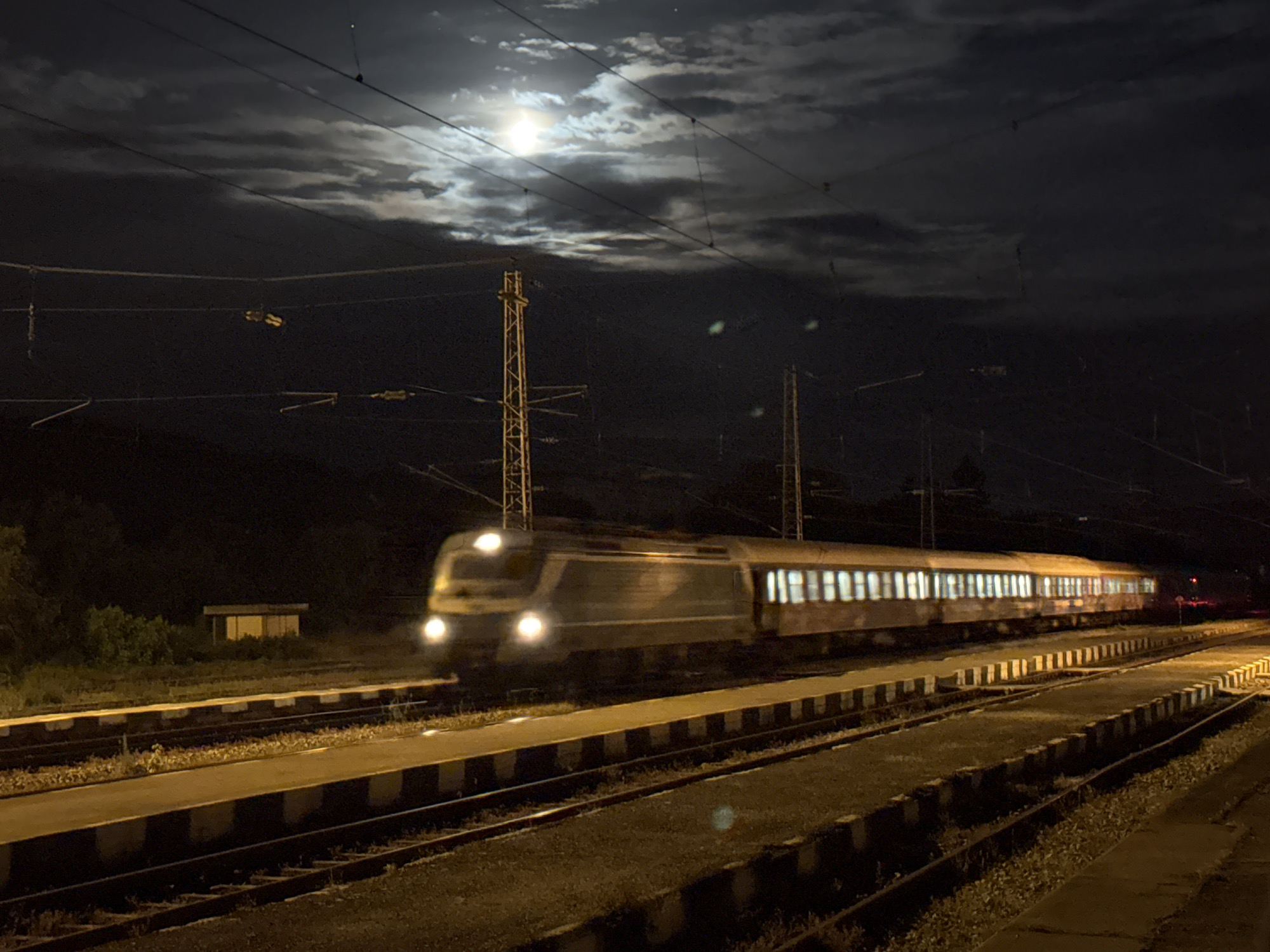 The image depicts a night scene at a railway station, specifically near the town of Strazhitsa, Bulgaria, based on the presence of Bulgarian rail infrastructure. A passenger train is in motion, its lights blurred due to the long exposure photography. The train is composed of multiple cars, each with illuminated windows. The sky is cloudy with a bright moon partially obscured by the cloud cover. Overhead electrical lines and supporting pylons are visible. The train tracks are lined with concrete sleepers, and the ground is covered in gravel. A small building is located to the left of the tracks. The scene is illuminated by the train's headlights, the moon, and other artificial lights.