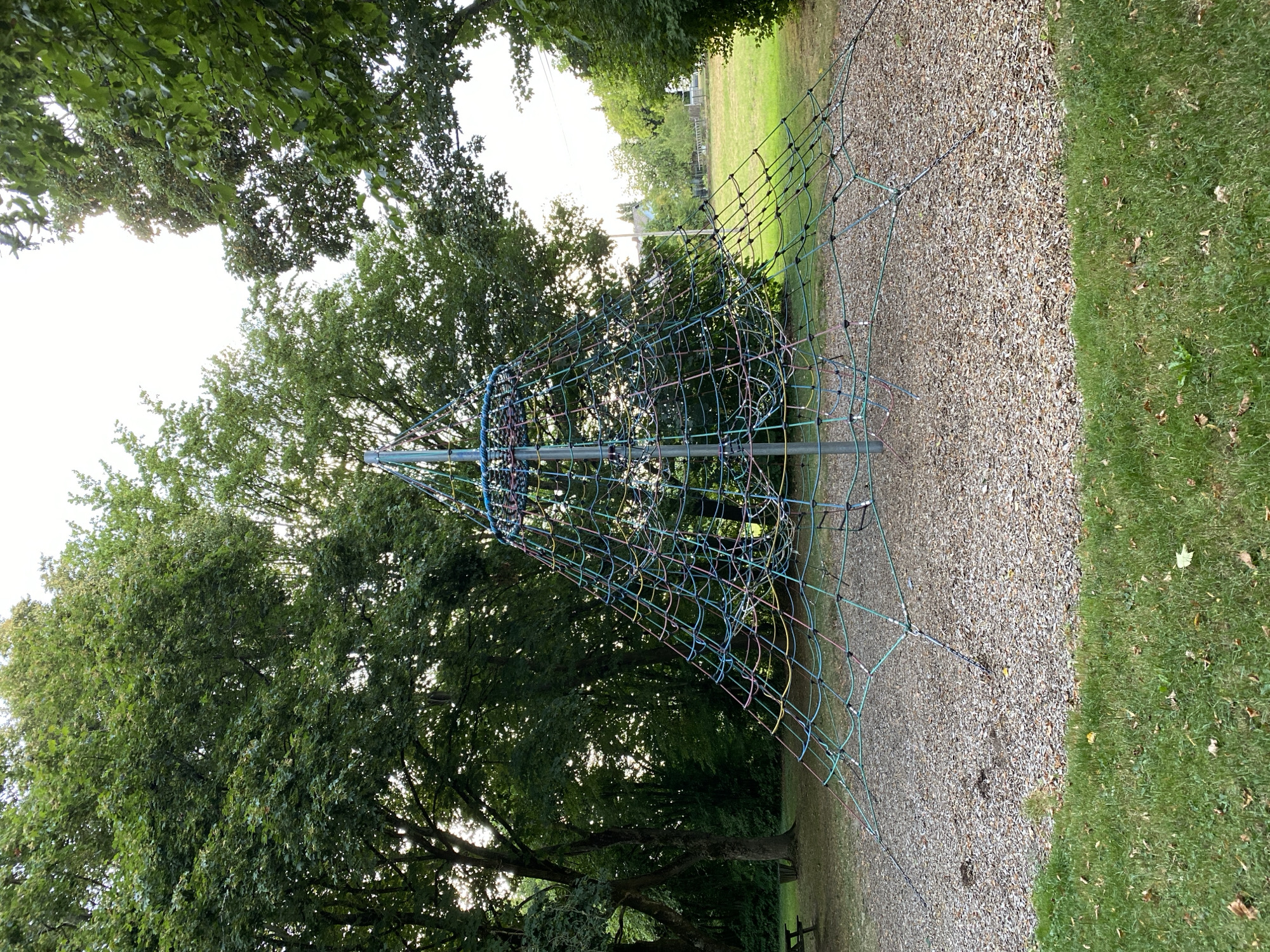 The image captures a pyramid-shaped climbing net structure situated in a park area. The structure is composed of various colored ropes, forming a dense net that expands outwards from a central pole. The ground beneath the climbing net is covered in wood chips, indicating a designated play area. Surrounding the play area is lush green grass. In the background, several mature trees with dense green foliage create a natural canopy. Further in the distance, a residential area with houses and a street is visible, suggesting the park is located within or on the outskirts of a town. The sky is bright and appears to be daytime. A picnic table is partially visible to the left of the climbing structure. The scene is static and shows no people or ongoing activity. The overall impression is one of a tranquil park setting, likely in Urbach, Germany, given the context provided.