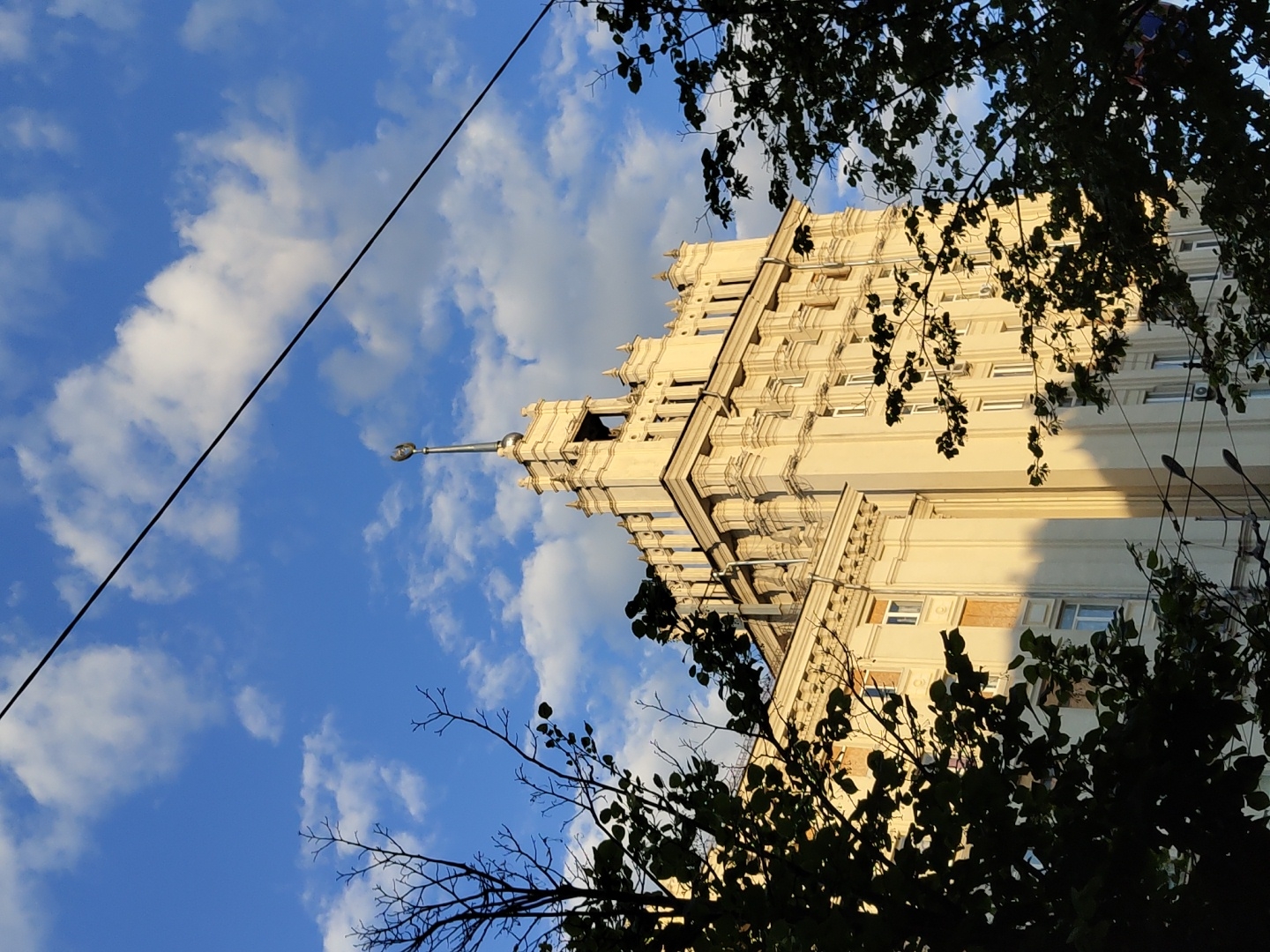 The image displays a towering, ornate building against a vibrant blue sky dotted with fluffy white clouds. The building, which appears to be a skyscraper or a grand residential block, dominates the left side of the frame and exhibits a cream or light beige facade. It features numerous classical architectural elements, including decorative cornices, pilasters, and recessed windows, suggesting a monumental style, possibly from the Soviet era.
A prominent, slender spire, topped with a finial, extends from the peak of the building, reaching towards the upper center of the photograph. Sunlight illuminates the building from an angle, creating strong contrasts and casting distinct shadows across its intricate details, indicative of either early morning or late afternoon.
A thick, dark utility cable or wire diagonally traverses the sky from the lower-left to the mid-right of the composition. In the foreground, dark, silhouetted tree branches and dense foliage partially frame the bottom-right and a small portion of the top-right of the image, adding depth and a natural contrast to the man-made structure.
The scene depicts an urban architectural landscape. There are no people visible, and no specific activity or event is taking place. The weather appears clear and sunny. No visible text is present in the image.