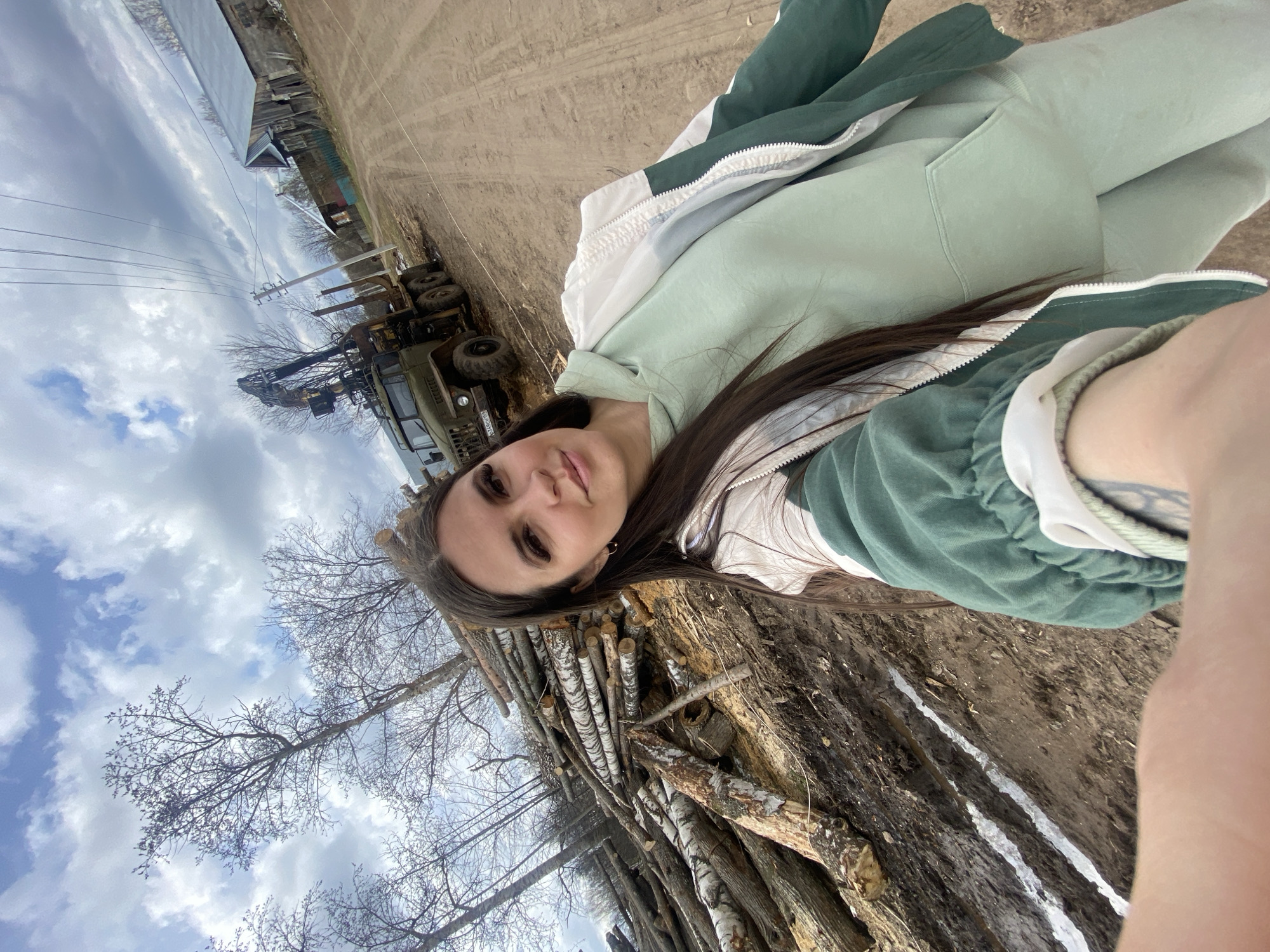 The image shows a woman taking a selfie. The background includes a stack of logs and a dirt road, with a utility vehicle and buildings visible in the distance. The sky is cloudy. The location is Zhigalki, Russia.