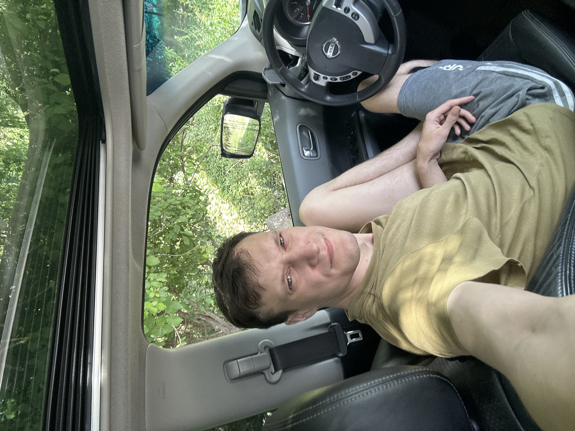 A man is pictured inside a car, sitting in the driver's seat. The car's sunroof is open, revealing a canopy of green trees and foliage overhead. The man is wearing a khaki-colored t-shirt and grey athletic shorts. His right arm is resting on his leg, and his left hand is visible on his lap. The interior of the car shows a black leather seat, a seatbelt, a steering wheel with a Nissan logo, and a side mirror reflecting the green surroundings. The backdrop outside the car is lush greenery, suggesting a rural or natural setting. The location is identified as Makiv, Ukraine.