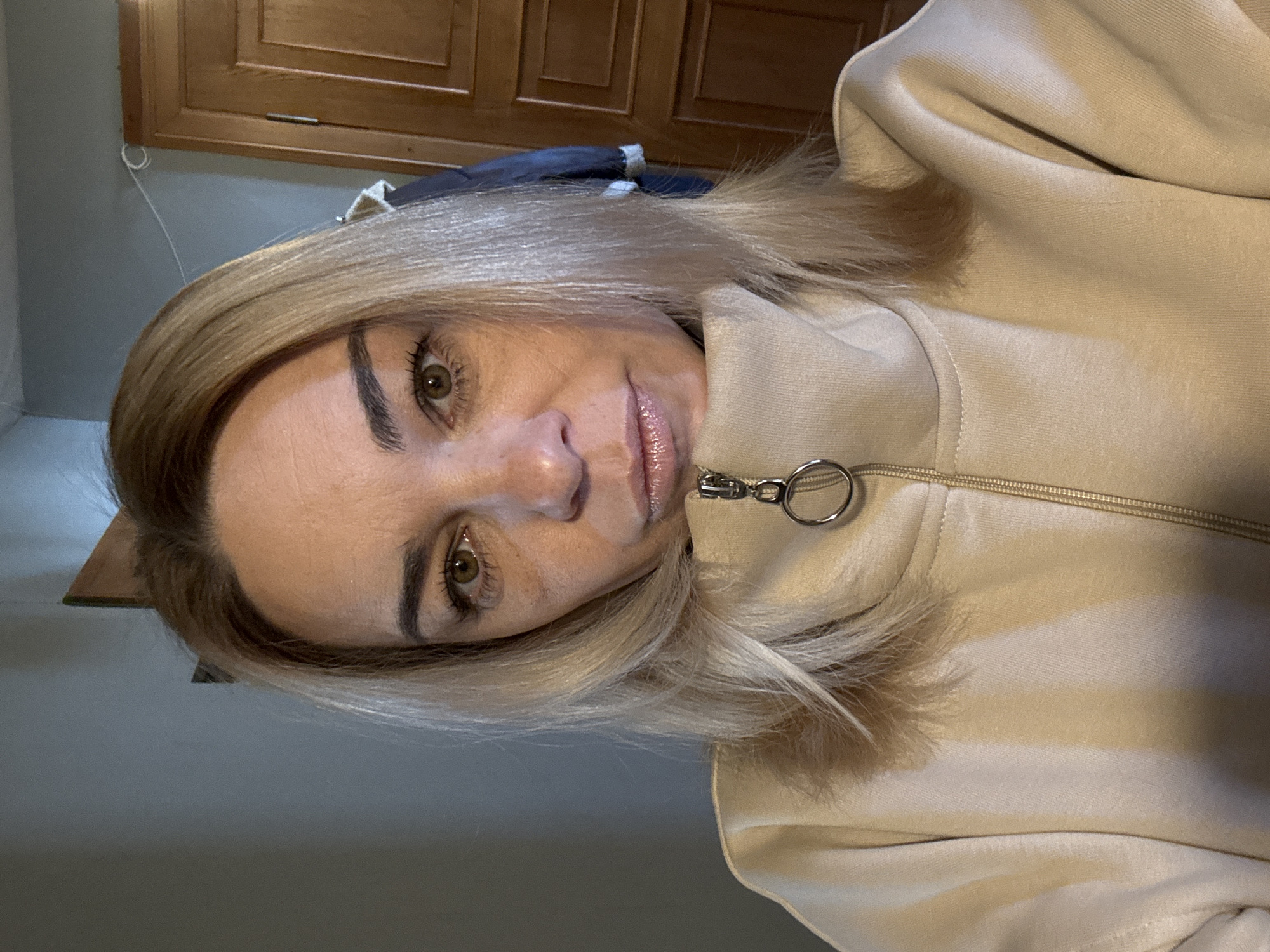 A close-up, eye-level shot of a woman with blonde hair and fair skin. She is wearing a beige jacket with a zipper, and the lighting suggests an indoor setting. Behind her, there is a wooden cabinet or wardrobe. No information suggests this photo was taken in Unknown, Russia.