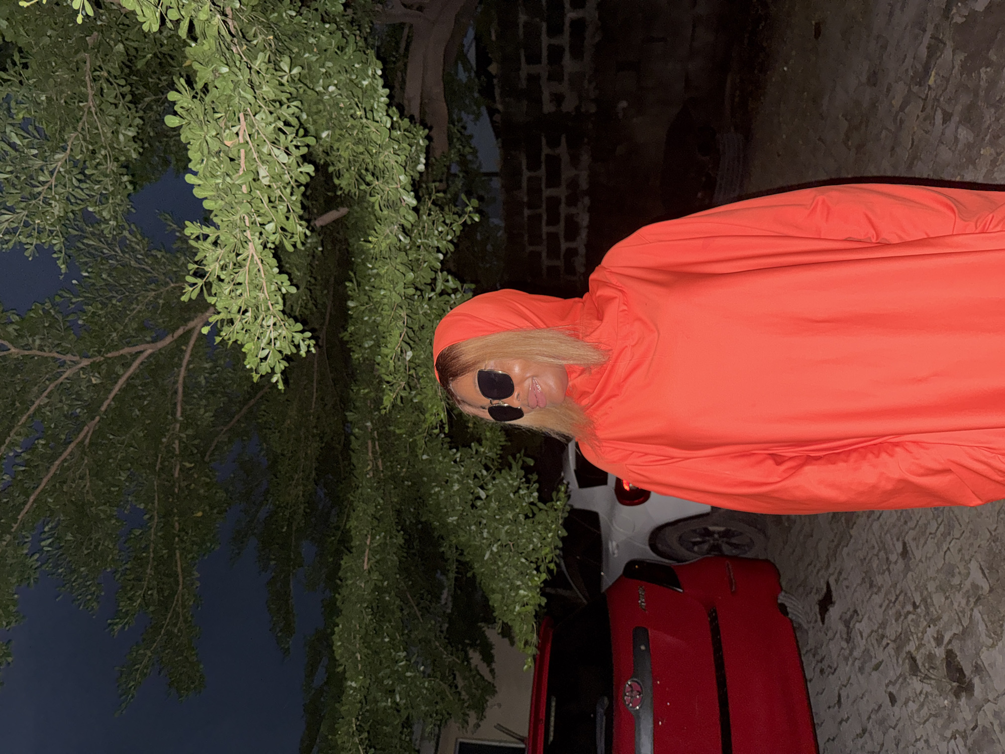 A woman with blonde hair and sunglasses stands outdoors at night, wearing an orange hooded garment. She is positioned in front of a tree with green leaves, and behind her, a red car and a white car are visible. The ground is paved with what appears to be interlocking stones. In the background, a brick wall can be seen. The scene is set in Ado, Nigeria.