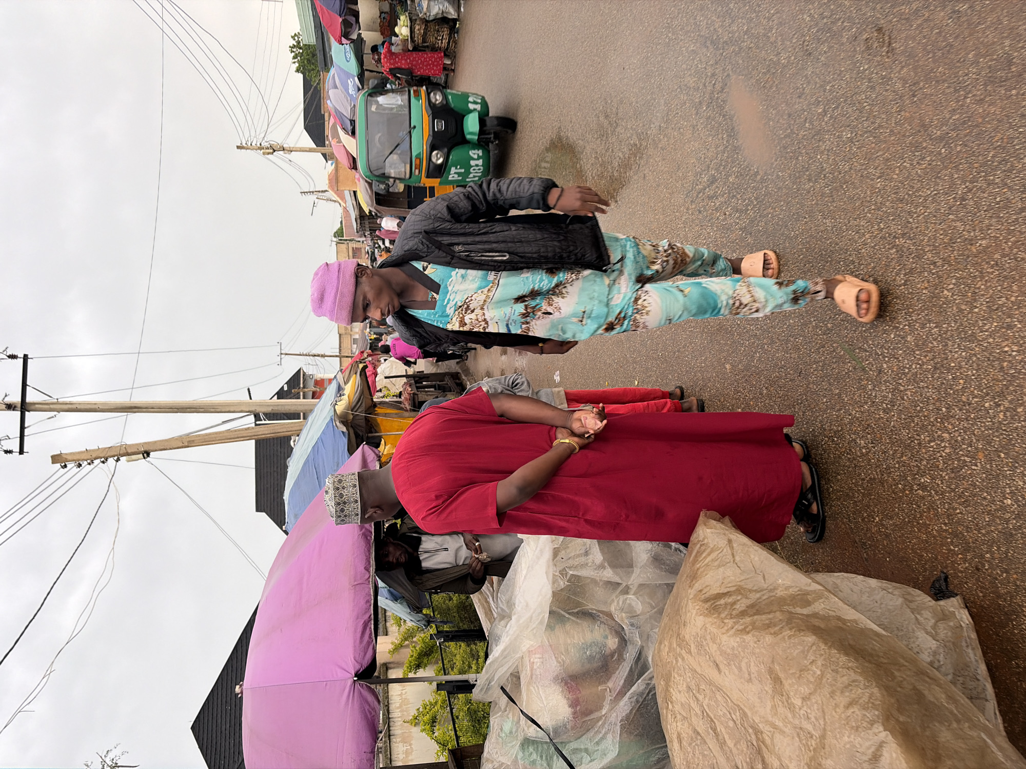 The image depicts a street scene in Jos, Nigeria, likely a market area under an overcast sky. In the foreground, two men are walking down a wet asphalt road. The man on the left, facing away from the camera, is wearing a vibrant red kaftan and a patterned kufi hat. His hands are clasped behind his back. He appears to be observing something to his left. To his left are large, transparently wrapped bundles of goods, possibly textiles or food items, and a large brown, crinkled-paper covered bundle.
The man on the right, walking towards the camera and slightly to the right, wears a black jacket over a light blue, patterned tunic with images of a beach and palm trees, and matching patterned trousers. He is also wearing a pink kufi hat and light brown slip-on sandals. His expression is neutral as he looks forward.
In the middle ground, a green and yellow three-wheeled auto-rickshaw (keke napep) with the license plate "PT-17814" is visible. Behind the auto-rickshaw, several market stalls with colorful umbrellas are set up. People are seen browsing and interacting. To the far left, under a large pink umbrella, another man in a dark jacket is partially visible. Further back, market stalls line the street, with some displaying goods under umbrellas of various colors, including blue and red. The environment suggests a bustling urban market scene, possibly after a rain due to the wet road surface. The overall atmosphere is busy and typical of a roadside market in Jos.