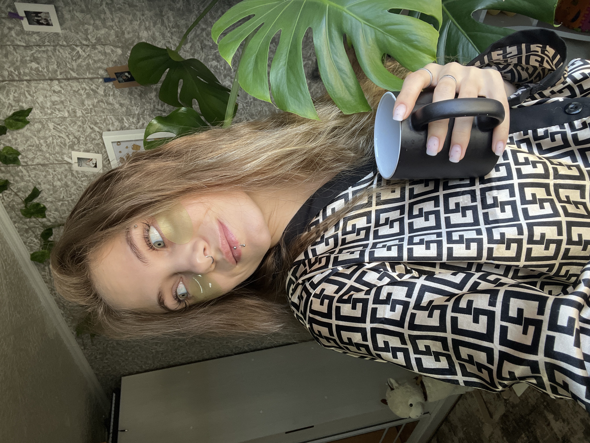 A young woman is lying on her back, holding a black mug with a white interior. She has blond hair and is wearing gold eye patches. She has a nose ring and a lip piercing. The woman is wearing a black and white patterned shirt. Behind her is a Monstera plant. A wall with pictures on it is visible in the background. A white stuffed animal is present on the lower right. The location Фоки, Russia is not discernable from the image.