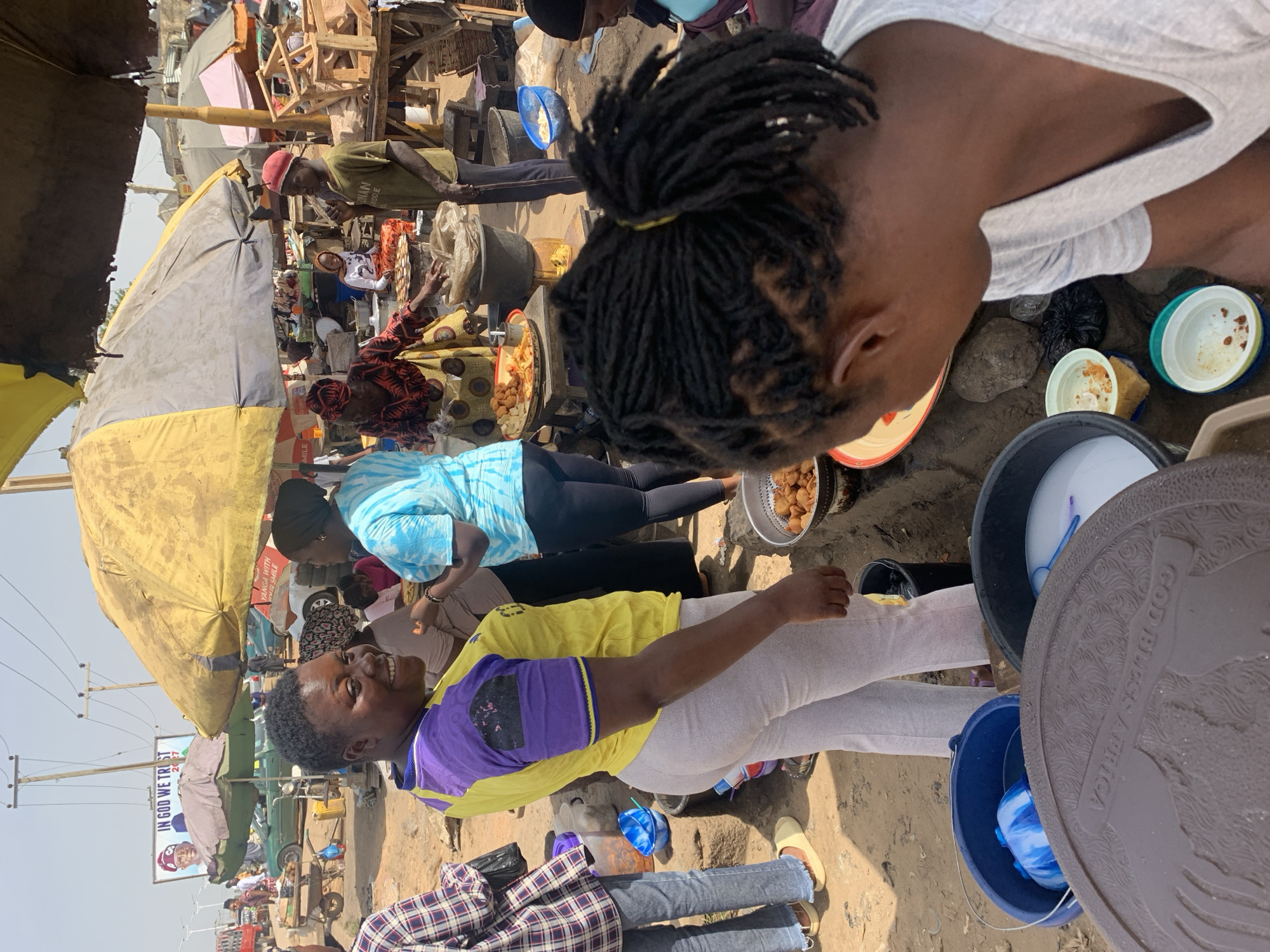 In the city of Rafin Par, Nigeria, a bustling outdoor market scene unfolds. Several people are gathered, with two women being prominent. One woman wears a yellow shirt with purple sleeves and light-colored pants, while the other, closer to the camera, wears a light-colored shirt and has her hair styled in dreadlocks. They appear to be engaged in some sort of food preparation or sales activity.
Around them, other individuals are visible, some under the shade of large, yellow umbrellas. Cooking pots, food items, and buckets are scattered around, indicating a market setting. A sign in the background reads "IN GOD WE TRUST". The overall environment is crowded and active.