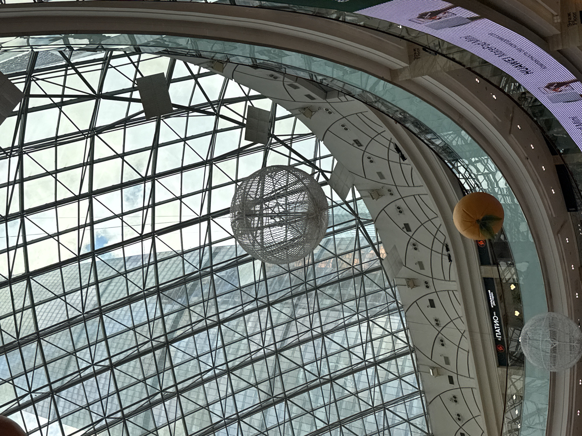 The image captures an upward view of the interior of the MEGA Khimki shopping mall in Khimki, Russia. A large glass dome structure with a grid-like metal framework forms the roof. Outside the dome, the sky is visible along with the tops of nearby buildings.
Suspended from the ceiling are several decorative spherical objects, including a gray, cage-like sphere and a yellow sphere resembling a fruit. A multi-tiered architectural element circles below the dome, with white surfaces accented by dark lines and shapes. On one level, signage with text in Russian is visible. Above, a digital display board runs along the circumference, showing advertisements. The lighting appears to be a combination of natural light from the dome and artificial lighting within the mall.