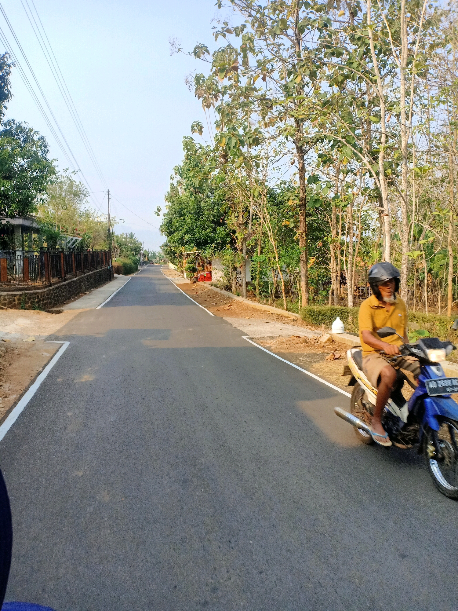 In the city of Jatinom, Indonesia, a man wearing a yellow polo shirt and brown shorts, along with a black helmet, rides a blue and white motorcycle down a paved road. The man is visible from the waist up, with his face partially obscured by the helmet's visor. He is positioned on the right side of the road, with the motorcycle's headlights on. The road is bordered by a dirt shoulder on the right, with sparse trees and vegetation. On the left side of the road, there is a low stone wall with a black fence, followed by more vegetation and houses. The sky above is a pale blue, suggesting it is daytime. The overall scene depicts a rural or suburban road in Indonesia with a single motorcyclist.