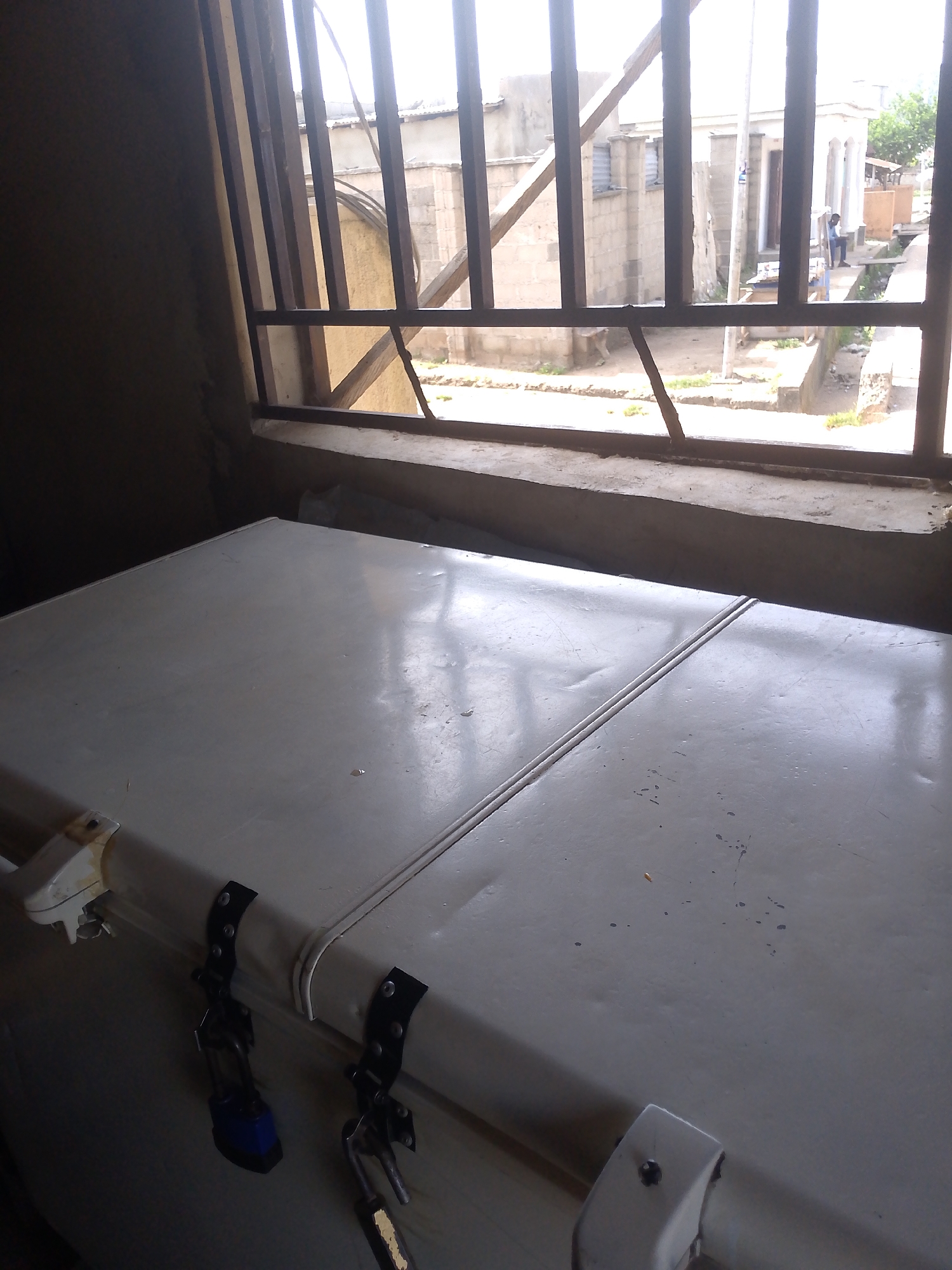A white chest freezer sits in the foreground, its lid slightly ajar, revealing a dark interior. Two black latches with attached padlocks secure the lid. The freezer is positioned in front of a window barred with metal grates. Outside the window, a street scene is visible, featuring buildings with rough concrete walls and corrugated metal roofs. A person is seated on a low wall to the right. The environment suggests an urban setting in Serti, Nigeria, possibly a commercial or residential area with basic infrastructure.