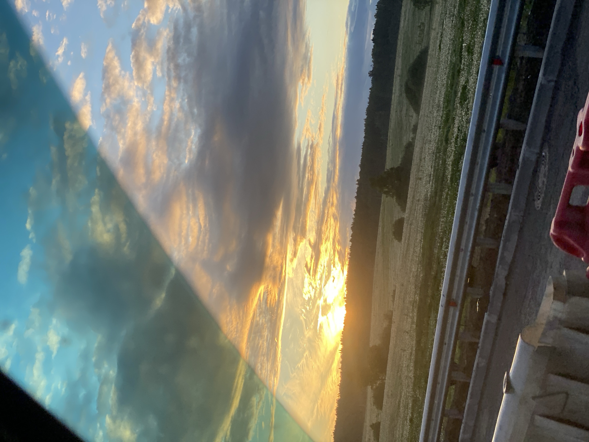 The image shows a landscape with a sky filled with clouds at sunset, reflecting in a car window. A field with trees is visible alongside a road with a guardrail and red traffic barrier. The location is in an unknown city, unknown.