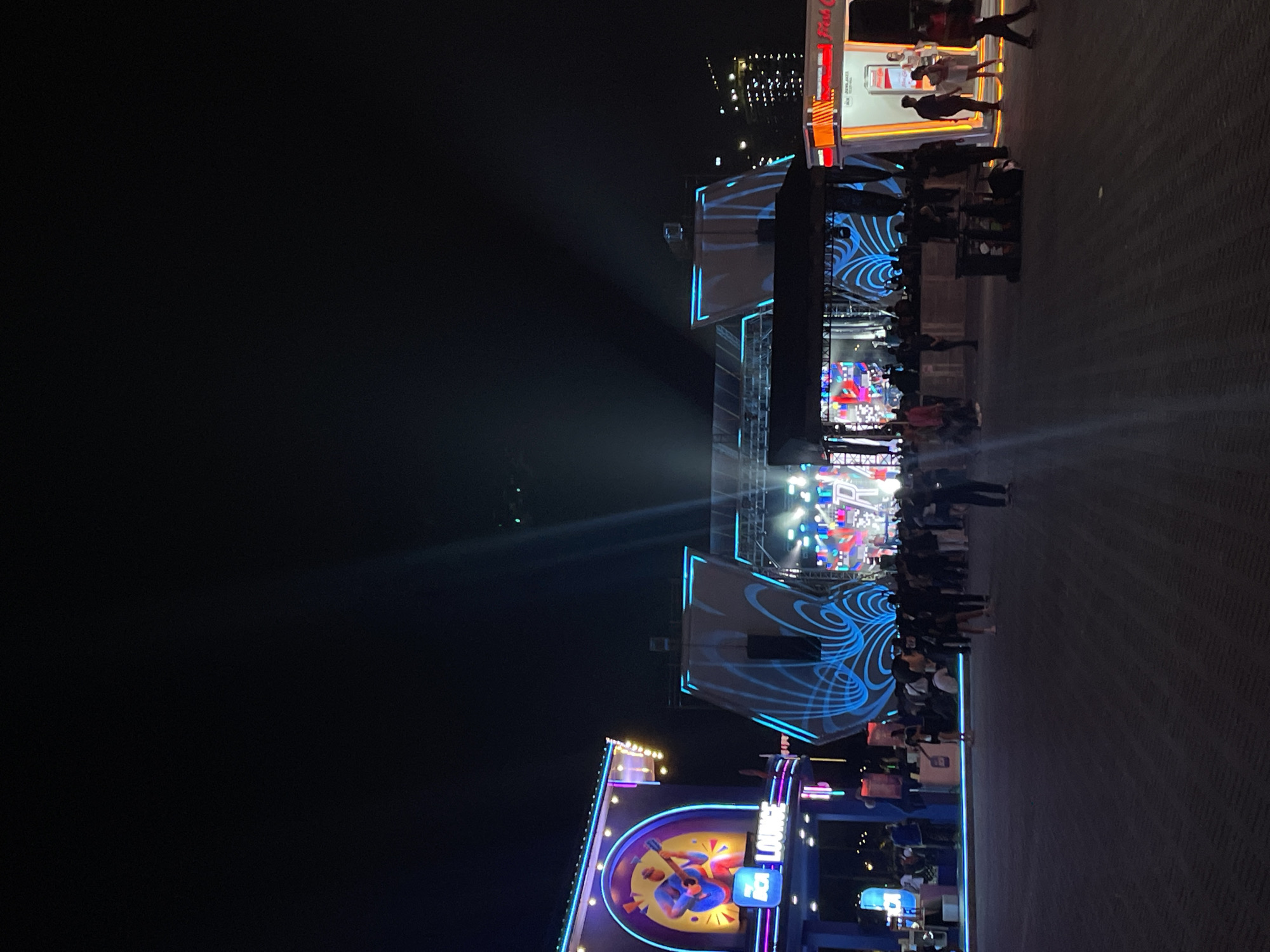 The image depicts an outdoor concert venue at night in Salembaran, Indonesia. A stage with a large screen displaying colorful visuals is centrally located. The stage is flanked by structures with blue neon designs. A crowd of people is gathered in front of the stage, facing it. Further back, structures with signage, including one mentioning "RCA Lounge," are illuminated. A Coca-Cola truck is visible in the background. The environment appears to be a paved area.