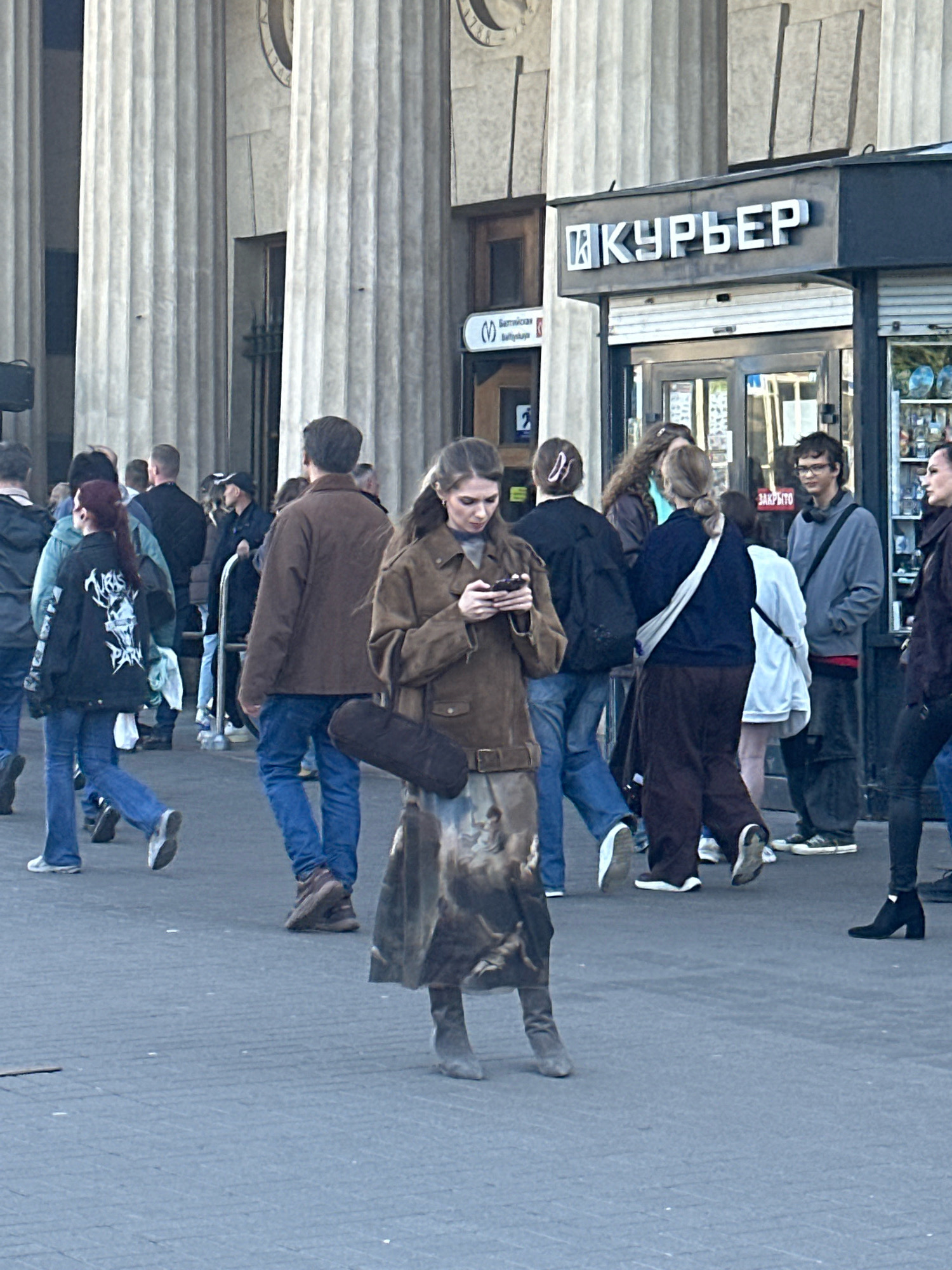 The image captures a street scene in front of a building with large classical columns, likely in Saint Petersburg, Russia, given the Cyrillic signage that reads "КУРЬЕР" (Courier). The focus is on a young woman in the foreground, wearing a brown trench coat and a patterned skirt, looking down at her smartphone. She has a dark brown cylindrical object, possibly a case for a musical instrument, slung over her shoulder. She is standing on a paved pedestrian area.
Behind her, a diverse group of people are walking and standing. To her left, a person with red hair is wearing a black jacket with a skull graphic. Further back, a man in a brown jacket is walking away from the camera, and another man in a grey hoodie is looking towards the camera. A storefront with a glass entrance and visible shelves of goods is to the right of the "КУРЬЕР" sign. The overall lighting suggests it is daytime.