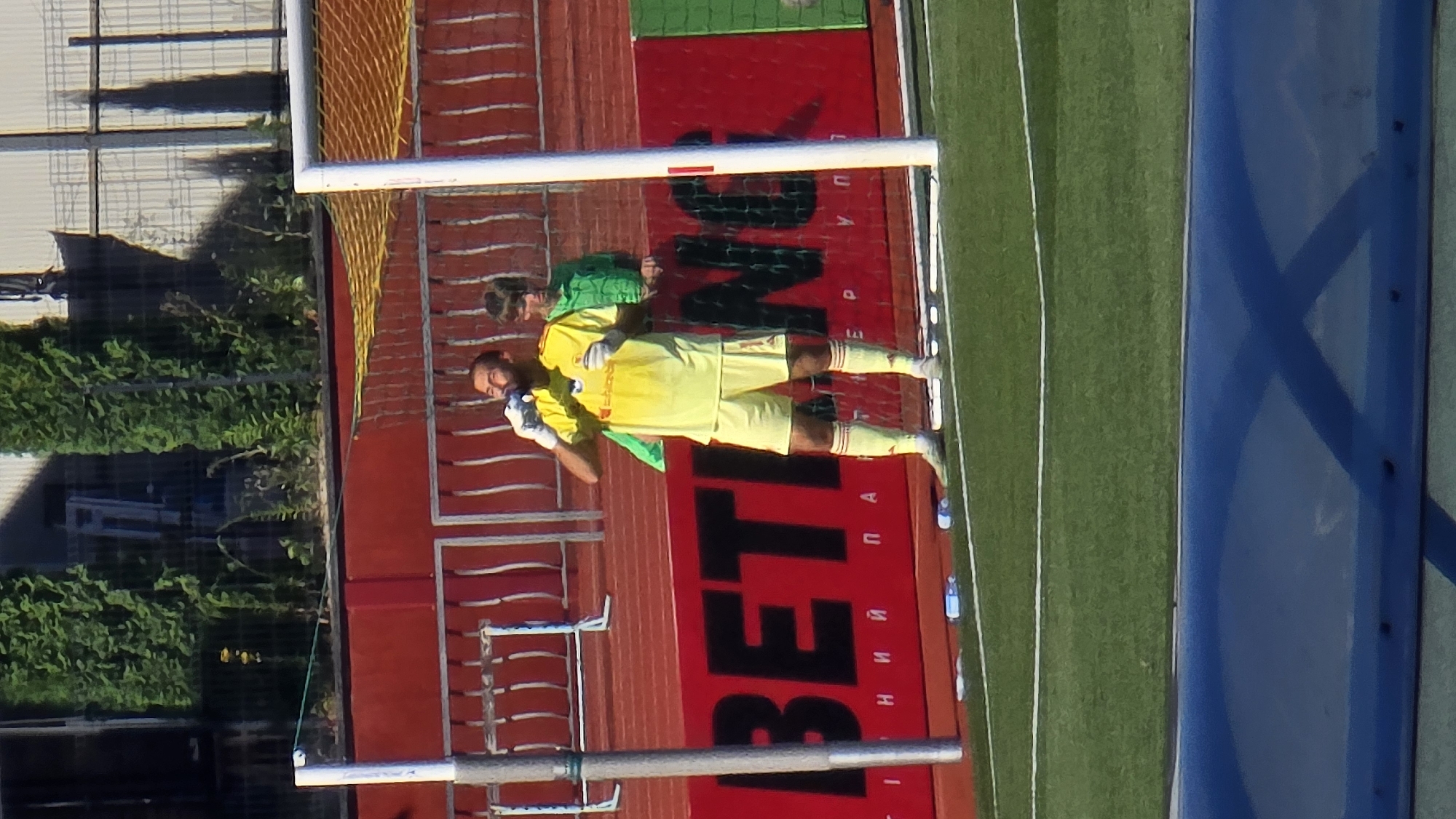 The image shows a soccer goalkeeper wearing a yellow jersey, yellow shorts, and red and white striped socks, drinking from a bottle near a goalpost on a sunny day. He is standing on a green soccer field with a red advertising banner in the background that reads "BETTING" in large black letters, along with some Cyrillic text. Another person in a green shirt is visible behind the goalkeeper, also near the goal. Several other water bottles are scattered on the field near the goalkeeper. The background includes a fence, trees, and a building. The scene appears to be at the Central City Stadium in Zhytomyr, Ukraine, a known venue for soccer matches.