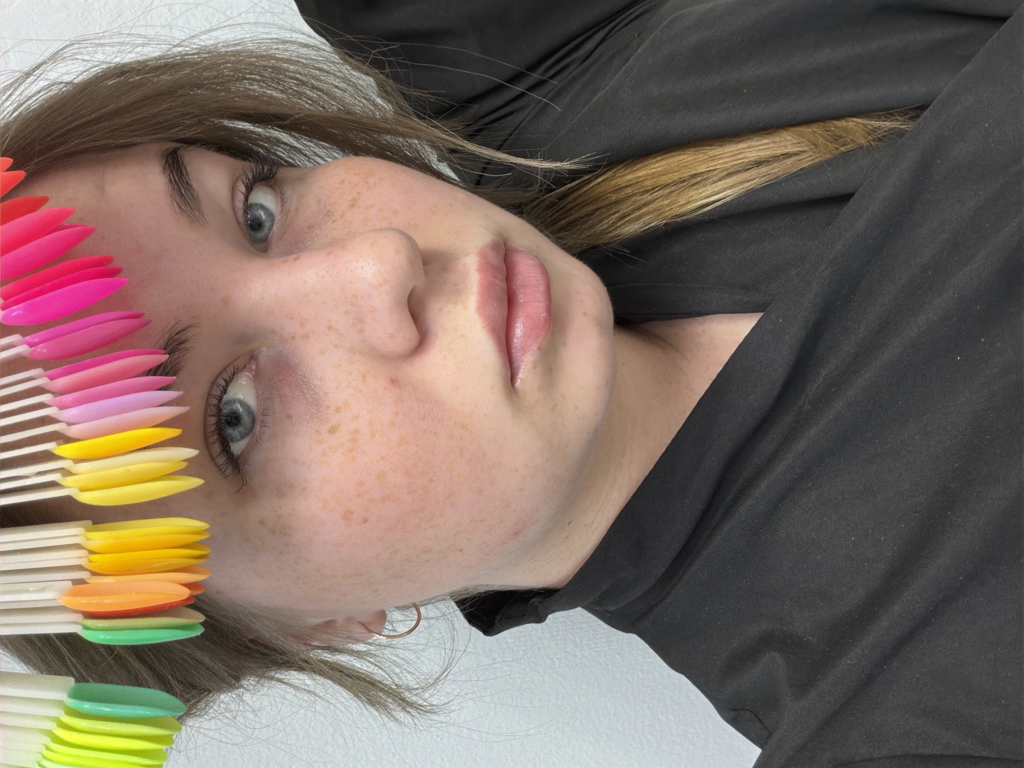 A young woman with light brown hair and blue eyes, wearing a black robe, poses for a selfie. She has fair skin with freckles and is wearing a small hoop earring in her left ear. Above her forehead, a set of colorful nail polish swatches are arranged in a semi-circle. The background is a plain white wall. The location in Chaikovsky, Russia cannot be determined from the image.