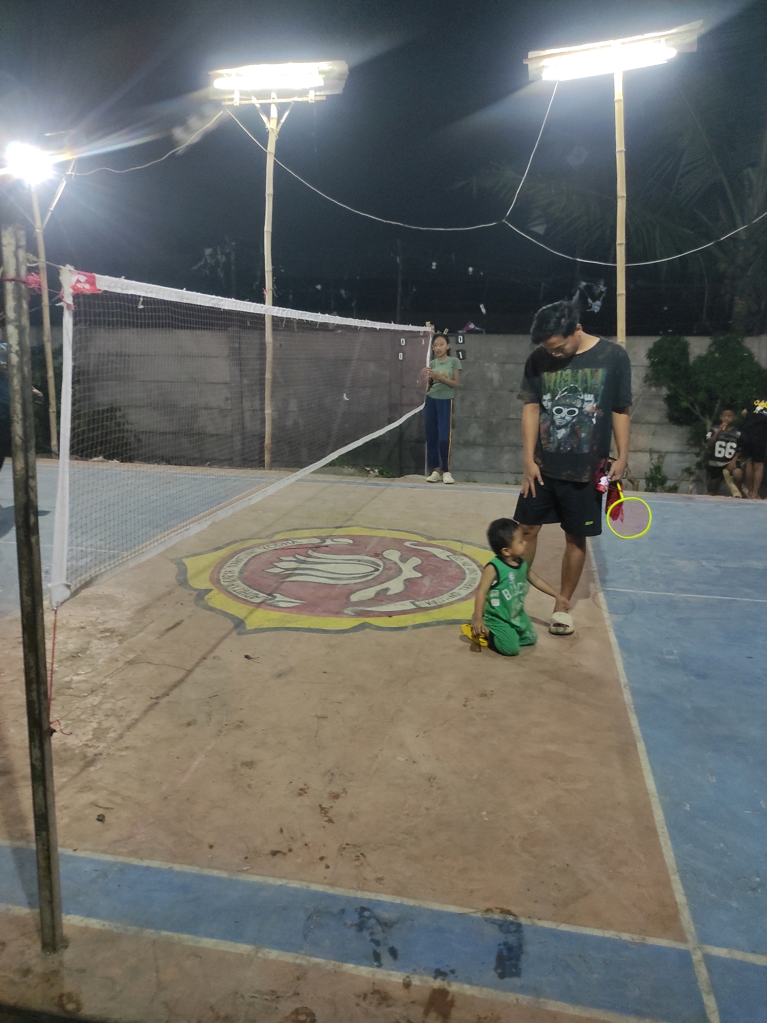 An outdoor badminton court in Padalarang, Indonesia, is illuminated by overhead fluorescent lights at night. A man wearing a black t-shirt with a graphic print and black shorts stands on the court, holding a yellow and pink badminton racket. Beside him, a young child in a green sleeveless shirt and shorts kneels on the ground. In the background, a young woman in a green t-shirt and blue pants stands near a scoreboard displaying "0 0" on one side and "0 1" on the other. Other individuals are visible further in the background. The court surface is a mix of concrete and blue paint, with a circular emblem featuring a bird design on the ground. A badminton net stretches across the court. The environment appears to be a casual sporting facility, possibly within a residential or community area in Padalarang.