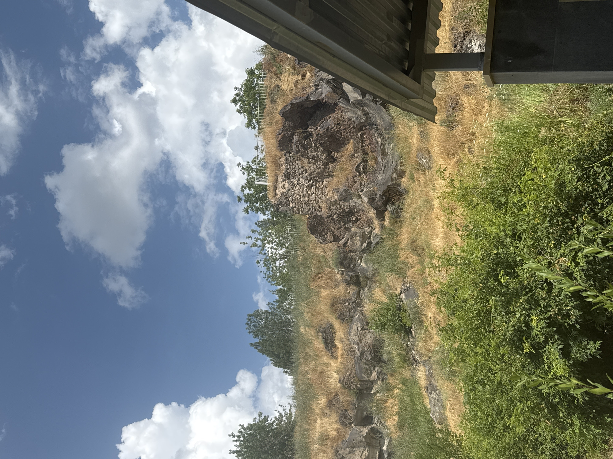 This image depicts a natural landscape with a partially visible man-made structure in the foreground. The background features a rocky hillside covered in dry, yellowed grass and scattered green bushes. Several trees are visible at the top of the hill, and some white metal fencing can be seen amongst them, suggesting a possible cemetery or memorial site. The sky is blue with scattered white clouds. The foreground shows the corrugated metal roof and a dark gray support pillar of what appears to be a covered pavilion or overhang. The overall scene is consistent with an outdoor environment, potentially in the vicinity of Malishka, Armenia, given the geographical context. There are no people or animals visible in the image.