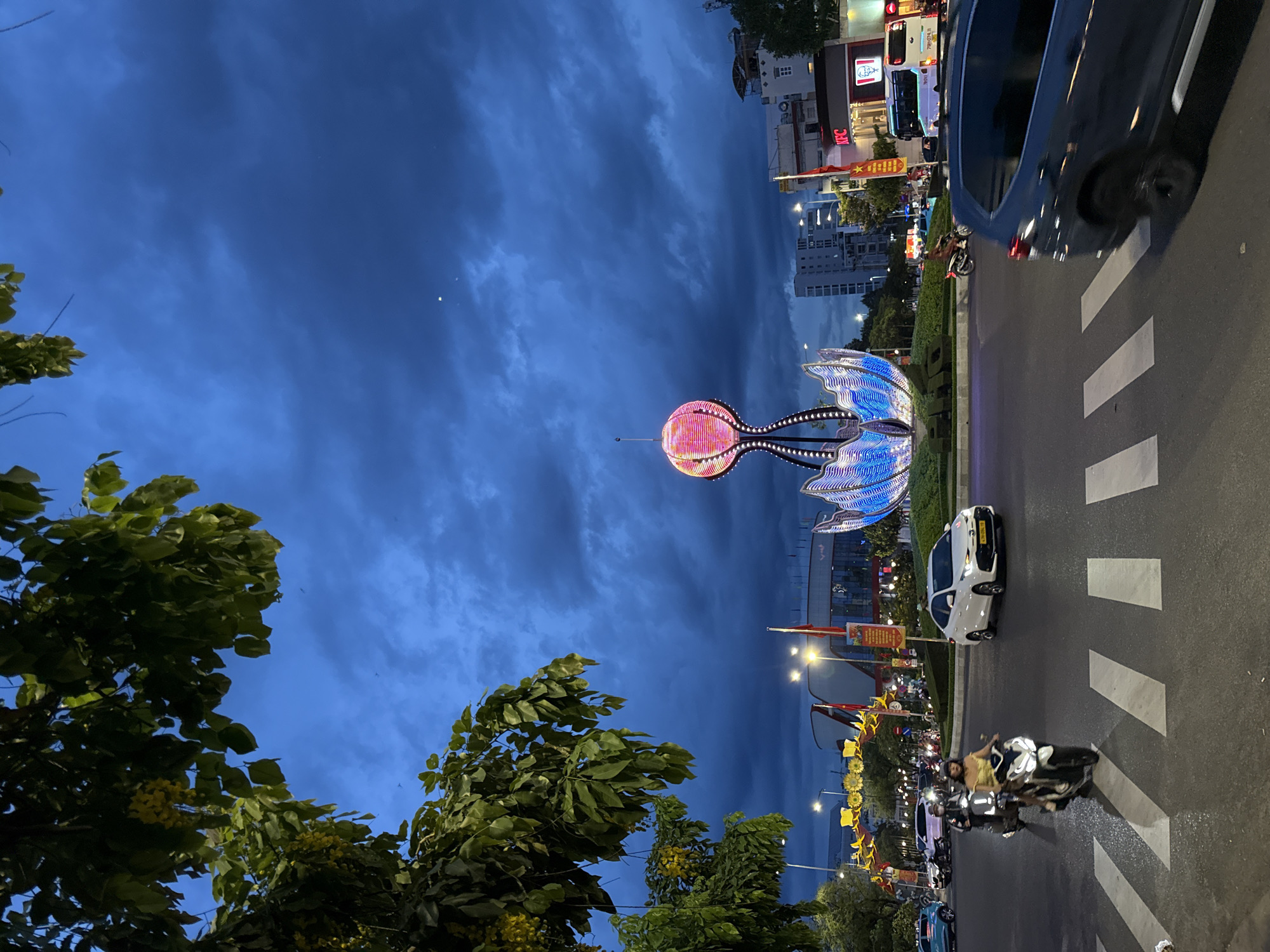 In Nha Trang, Vietnam, at dusk, a large, illuminated sculpture resembling a lotus flower stands in the center of a roundabout. The sculpture is adorned with colorful lights that transition between pink, orange, and blue. Surrounding the roundabout, streetlights and building lights illuminate the scene. Traffic flows through the roundabout, with several motorcycles and cars visible. On the right side of the frame, a KFC restaurant with its iconic Colonel Sanders logo is present, along with other businesses and buildings. Lush green trees with yellow flowers hang from the left side of the image, framing the sky, which is filled with dark, dramatic clouds. The overall atmosphere suggests a lively urban environment at the end of the day.