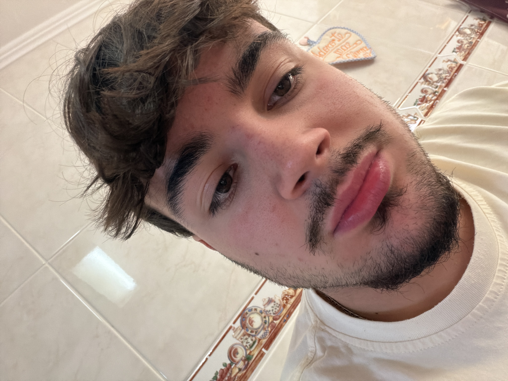 A close-up, selfie-style shot of a young man with dark hair and a beard. He is wearing a light-colored t-shirt. The background includes tiled surfaces and a decorative tile border with a floral pattern. A heart-shaped decorative object is visible in the background, with text on it which translates to "I dedicate to you". There is no explicit information about the location being Vila Real, Portugal, so I cannot include this in the description.