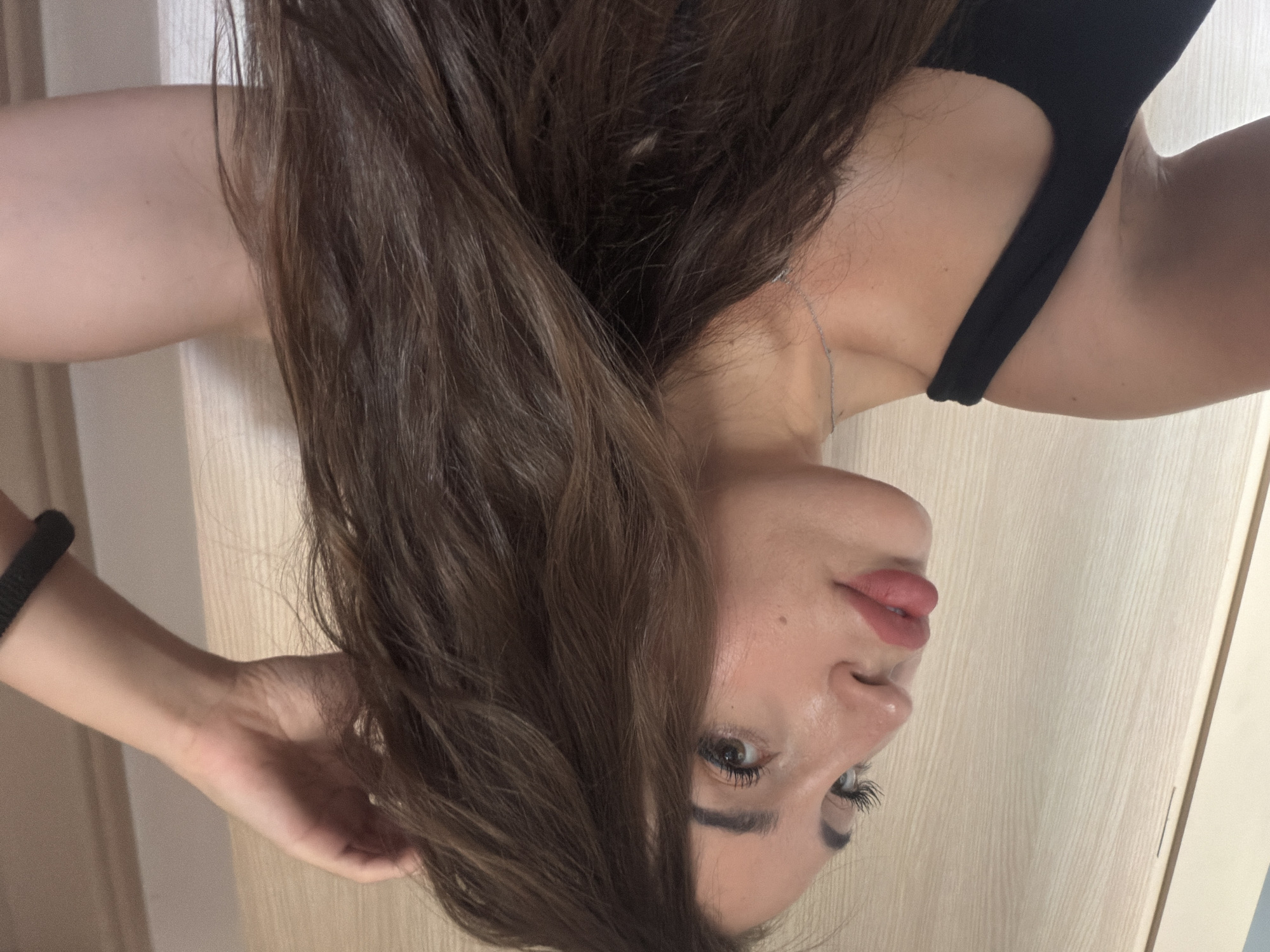 The image depicts a close-up, profile view of a young woman in Tersefanou, Cyprus. She has long, brown hair that is slightly messy, and she is touching the back of her head with her right hand. Her left arm is bent at the elbow and appears to be resting on her left side. She is wearing a black, sleeveless top and a thin silver necklace. Her makeup includes defined eyebrows, mascara, and a matte pink lipstick. The background consists of a pale wooden door or wall. The lighting suggests an indoor setting, possibly a home.