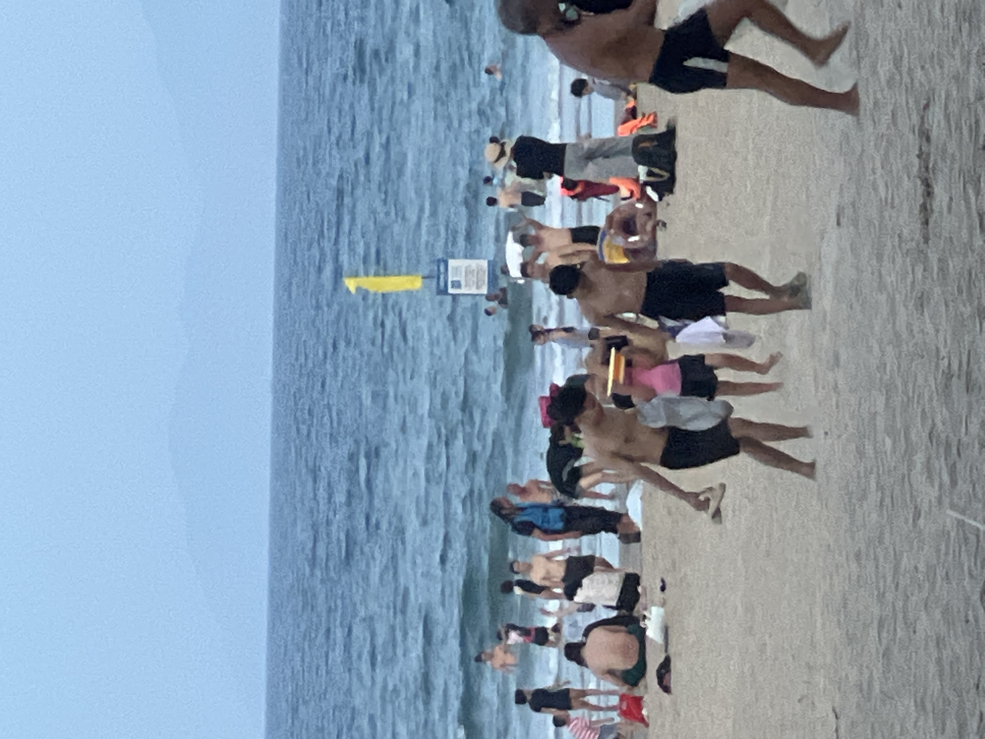 The image depicts a crowded beach scene at My Khe Beach in Da Nang, Vietnam. The sky is overcast with a soft, diffused light. The sea is a tranquil blue, with gentle waves rolling towards the shore. The beach itself is sandy, with numerous people engaging in various activities. Some are swimming or wading in the water, while others are relaxing on the sand. There are groups of people walking along the shoreline, some carrying towels or bags. A few individuals are playing with beach toys, such as a frisbee visible in the foreground. Beach umbrellas and towels are scattered across the sand, adding to the vibrant atmosphere. In the distance, a yellow flag and a blue sign are visible, possibly indicating safety information or designated swimming areas. The overall impression is one of a lively and enjoyable summer day at the beach.