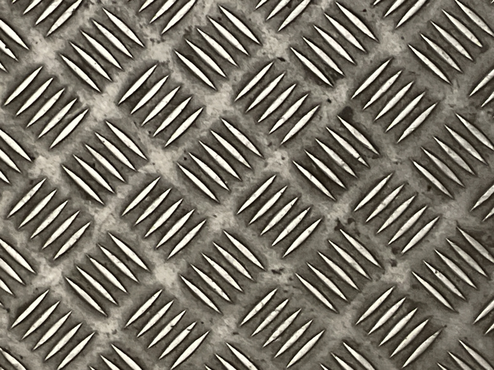 The image provides a close-up view of a metal diamond plate, also known as checker plate or tread plate. The primary subject is a textured metallic surface designed for anti-slip properties. It features a repeating pattern of raised, elongated oval or lozenge shapes, arranged diagonally in staggered rows across the entire frame.
The metal appears to be aged and worn, exhibiting a range of tones from bright silver on the highlights of the raised patterns to darker greys and muted, earthy browns in the recessed areas. These darker, mottled areas suggest grime, stains, or light corrosion on the base metal. The raised elements are brighter and reflect light, giving them a distinct three-dimensional appearance and highlighting the texture.
No specific activity or event is taking place, as the image is a static, detailed shot of a material surface. There is no visible text, and the setting is implied to be an industrial, commercial, or utilitarian environment such as a floor, ramp, or walkway, given the nature of diamond plate. The lighting is even, emphasizing the contrast and texture of the metal.