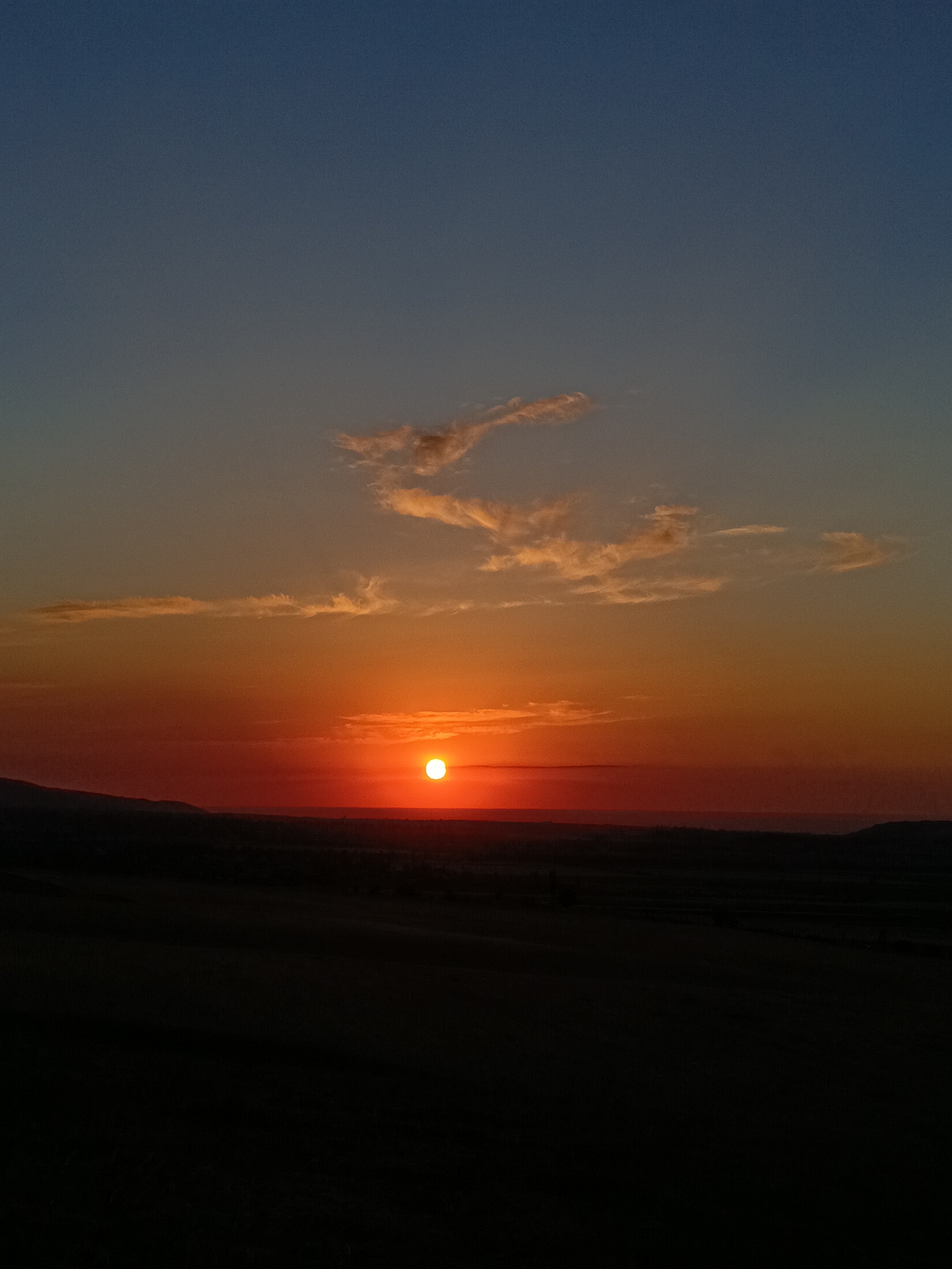 The image is a vertical photograph of a sunset over a rural landscape. The sky transitions from a deep blue at the top to a vibrant orange and red near the horizon. A small, bright orange sun is visible just above the horizon, casting a warm glow across the sky. wispy clouds are scattered across the upper half of the sky, catching the light and appearing in shades of white and orange. Below the sky, a dark, silhouetted landscape stretches across the frame. This appears to be rolling hills or mountains, with some suggestion of vegetation or fields in the lower portion of the image. The overall mood of the image is serene and peaceful, capturing the natural beauty of the end of the day. The location is Arashan, Kyrgyzstan.