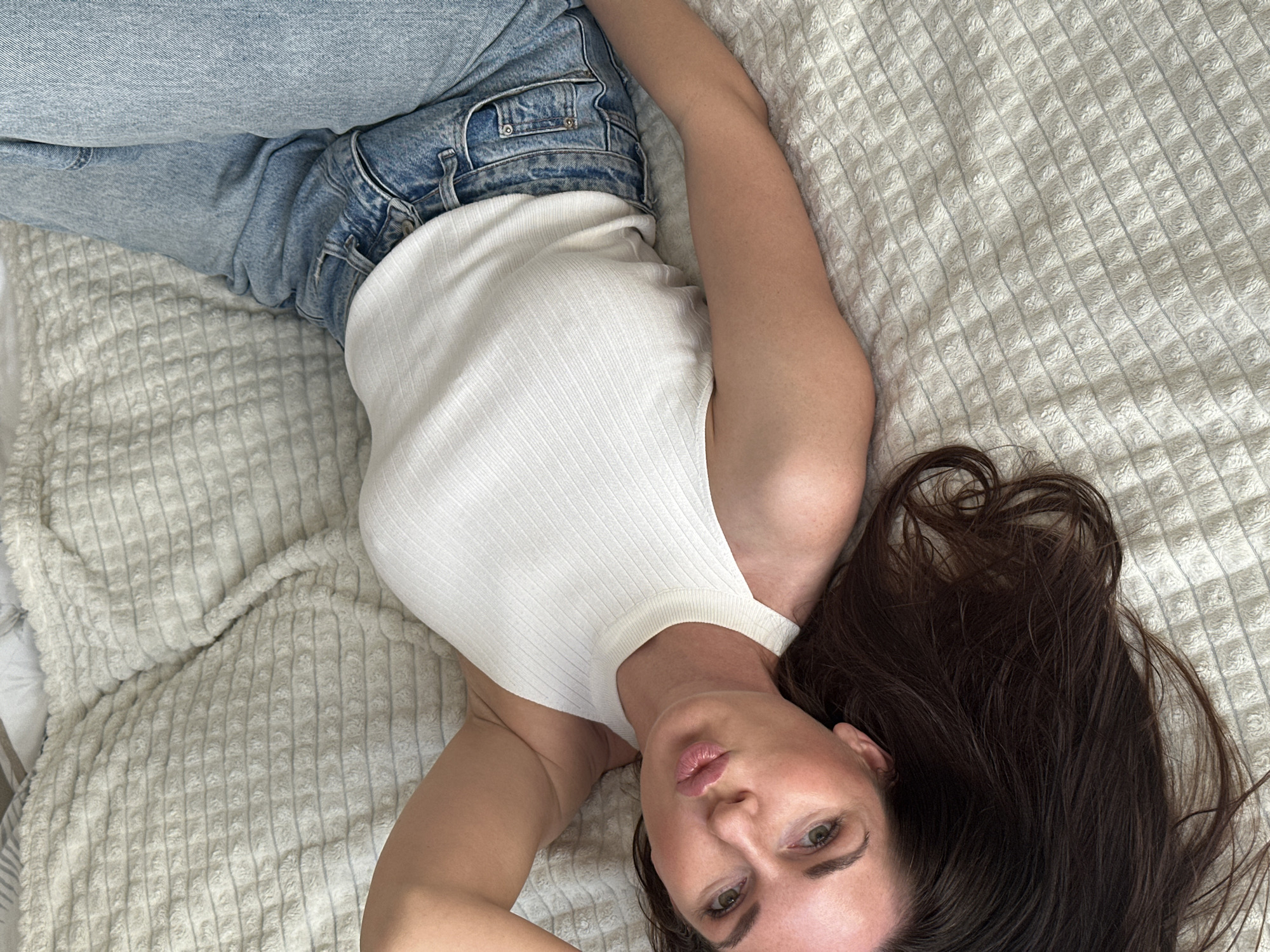 A woman is lying on a light-colored textured blanket. She is wearing light blue jeans and a white ribbed tank top. She is looking at the camera with pursed lips. Her long brown hair is spread out on the blanket.
There is no information in the image to confirm that this scene is in Chaikovsky, Russia.