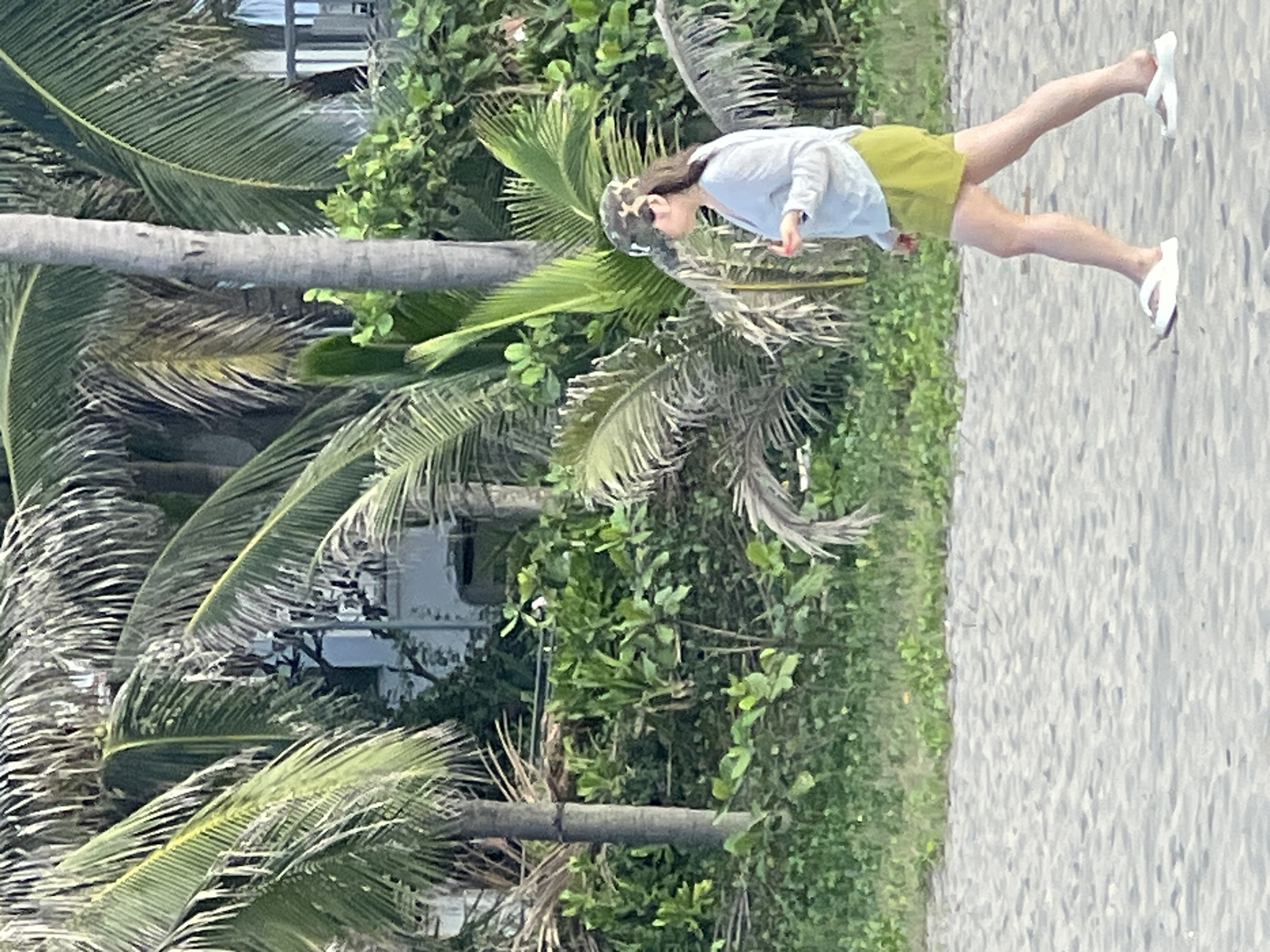 The image shows a person walking on a sandy beach in Đà Nẵng, Vietnam. The person is wearing a light blue long-sleeved shirt, lime green shorts, a camouflage-patterned baseball cap, and white platform sandals. They are walking from left to right in the frame, with their body angled away from the camera. Behind the beach, there is dense green foliage, including palm trees, and a building with a balcony railing is visible in the upper right corner. The sand in the foreground has ripples from the wind or water.