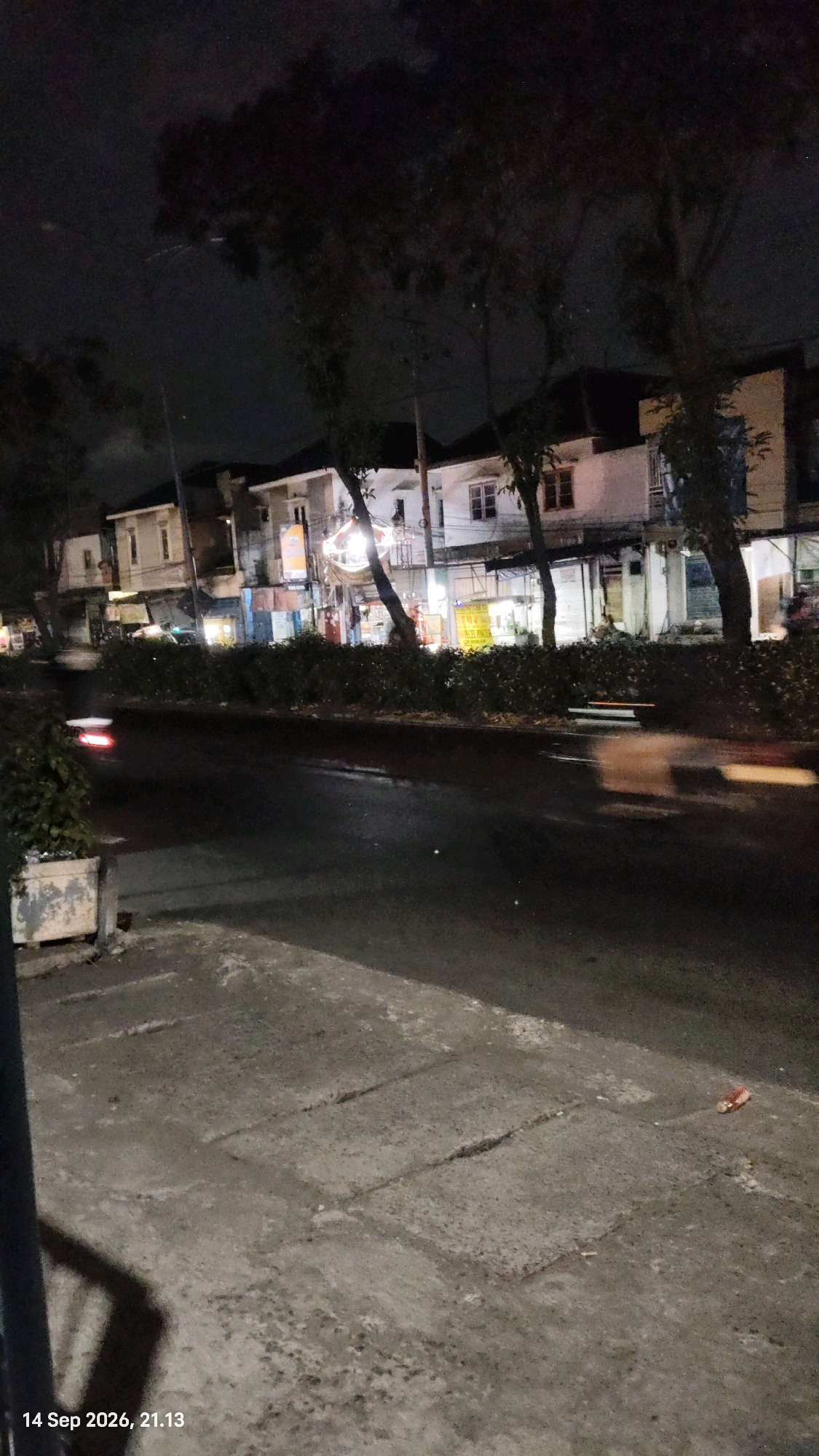 The image depicts a street scene at night in Surabaya, Indonesia, dated September 14, 2026, at 21:13. A two-lane road runs horizontally across the frame, with its surface appearing wet and reflective, suggesting recent rain or dew. On the left side of the road, a concrete planter box is visible, containing some sparse green foliage. The sidewalk, made of gray concrete slabs with visible cracks and repairs, occupies the lower foreground and left edge of the image.
Across the street, a row of multi-story buildings lines the road. These buildings appear to be commercial establishments, with some displaying illuminated signs. The architecture suggests a mix of older and newer construction. Trees with dark foliage are scattered throughout the scene, partially obscuring the buildings and the night sky. The sky itself is dark and overcast, with some faint, possibly artificial, light sources visible.
A vehicle with its headlights illuminated is in motion on the road, indicated by a blur of red light on the left side of the image. Another blurry light streak, possibly from another vehicle, is present on the right side of the road. The overall impression is of a quiet urban street after dark, likely in a residential or mixed-use area of Surabaya. The lighting is dim, with artificial lights from the buildings and the vehicle being the primary sources of illumination.