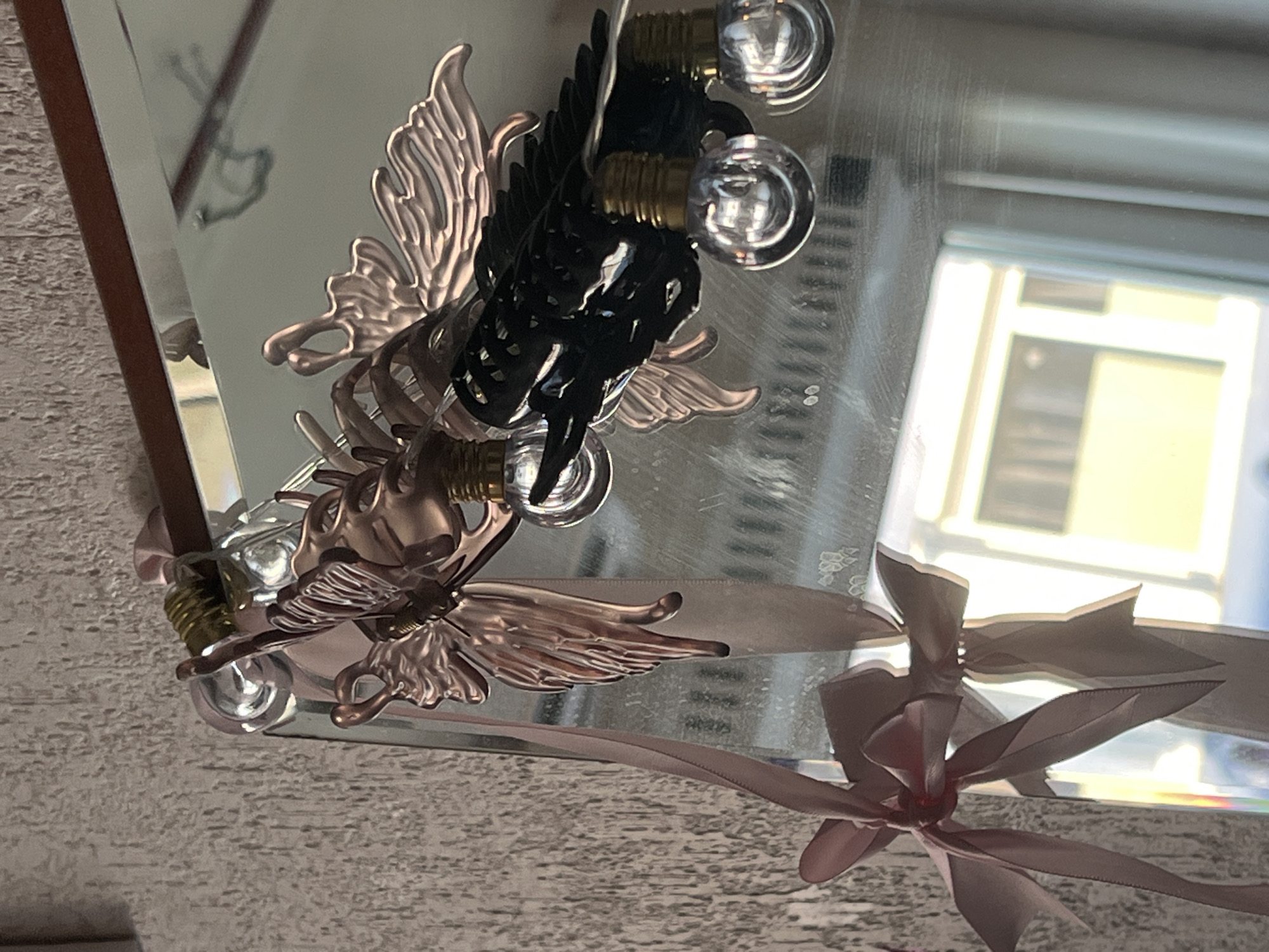 The image shows a close-up of a decorative mirror with various ornaments. A pink ribbon is tied in a bow to the left side of the mirror frame, with the ribbon extending downwards. Attached to the top of the mirror are metallic ornaments resembling insects or mythical creatures, with delicate wings and intricate details. Small, clear bulb lights are strung along these ornaments. The mirror reflects a room with textured walls and a window with a view of buildings. The location is not specified in the image or its context, therefore it cannot be determined to be Genemuiden, Netherlands.