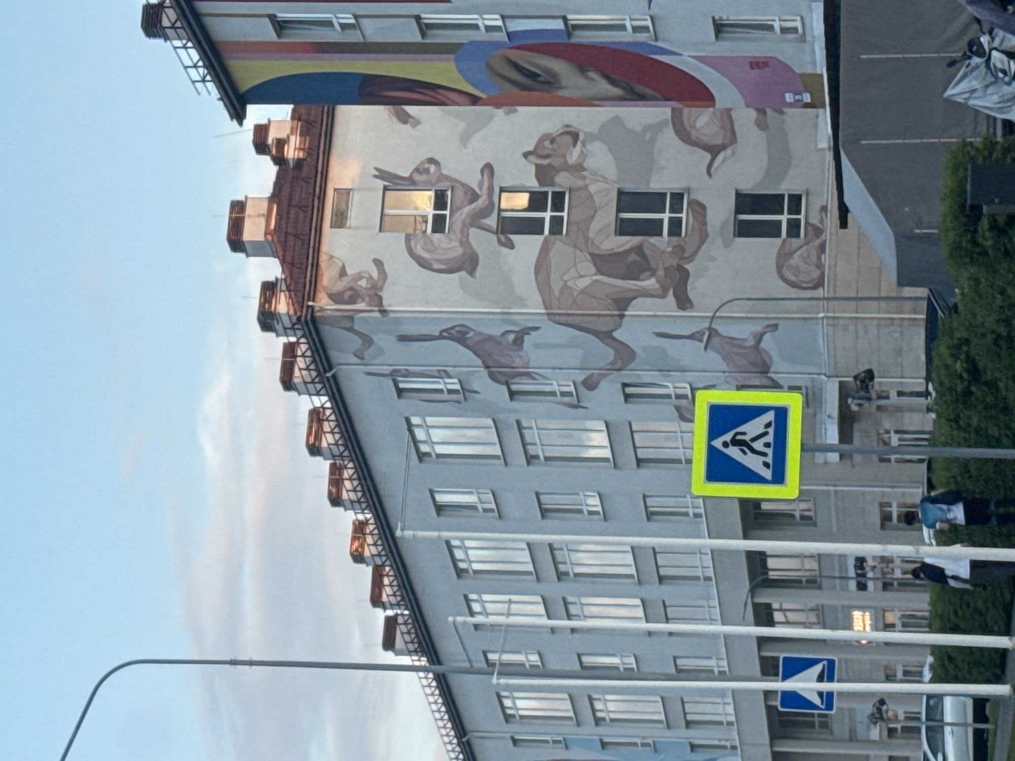 The image depicts a multi-story building with a mural on its side. The mural features abstract figures of animals. There is a pedestrian crossing sign in the foreground. People are walking near the building. The location is likely Sirius, Russia, based on the prompt.