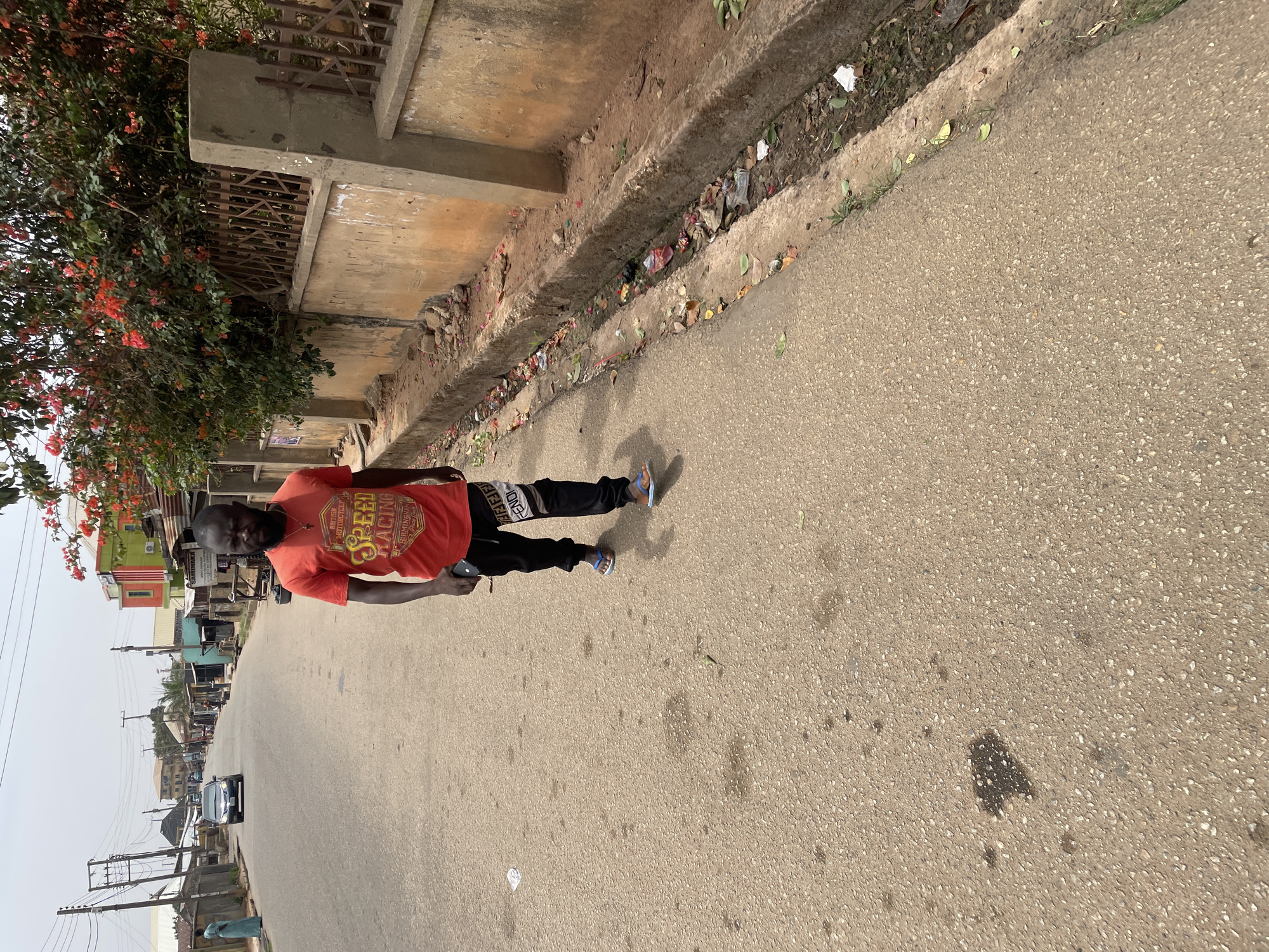 A man stands in the middle of a paved road. He is wearing an orange t-shirt with a graphic design and black pants with white lettering on the side. His feet are clad in blue sandals. The road is grey and shows signs of wear. To the left, there's a building and a sidewalk covered in litter and plants. In the distance, buildings line the road, and a car is driving towards the man. Power lines stretch across the sky. The photo appears to be taken in Jos, Nigeria, based on the style of the buildings, the type of foliage, and the general environment.