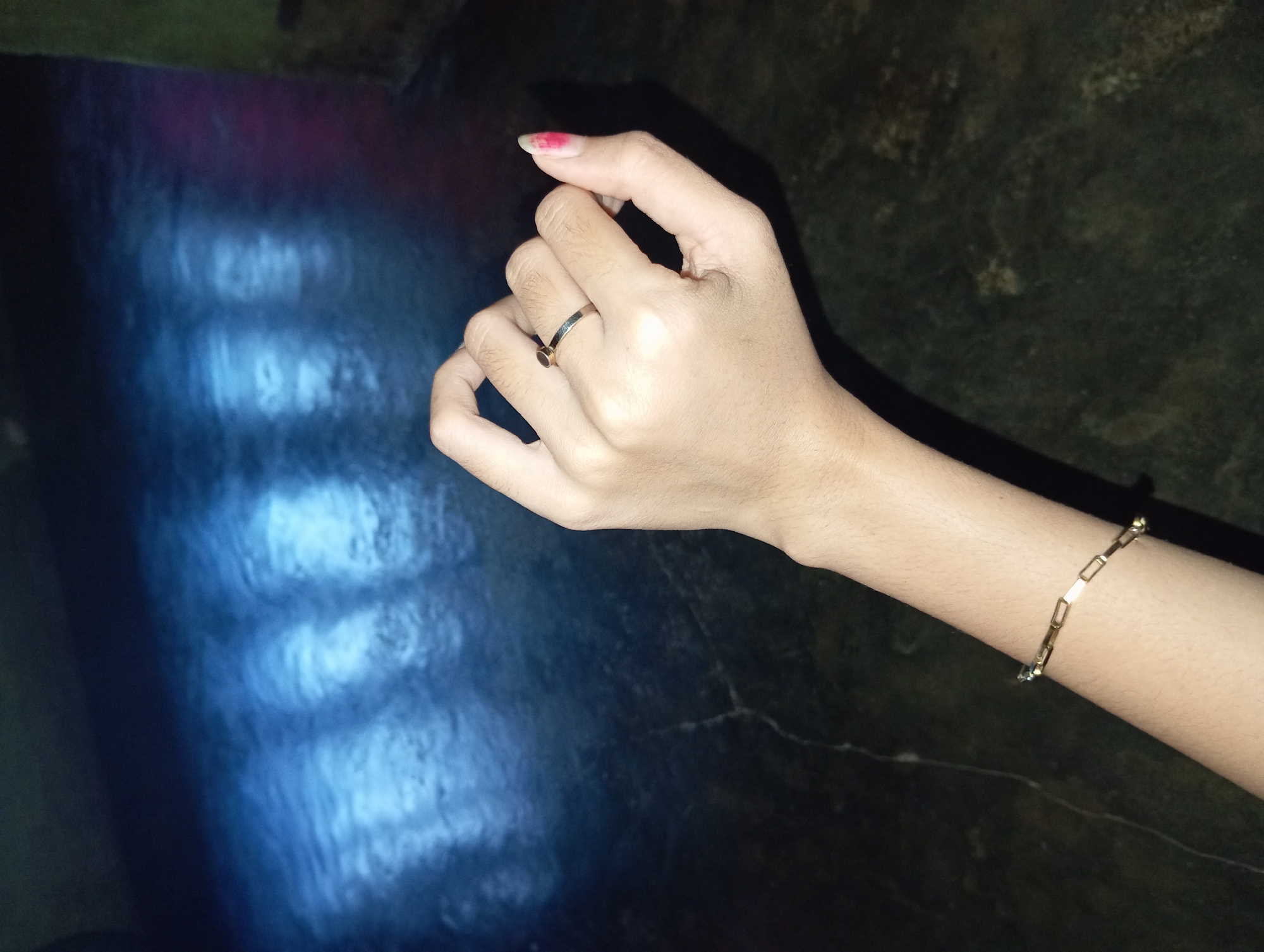 A close-up, low-angle shot shows a person's left hand and forearm against a dark, textured surface. The hand is positioned with the palm facing upwards, and the fingers are slightly curled. A thin, silver ring with a dark stone is worn on the ring finger. The pinky finger is extended, and the nail on this finger has a small amount of red polish. A gold-colored bracelet, made of interconnected rectangular links, is worn on the forearm. The background is dark and appears to be a wall or floor, with a distinct reflection of light and what looks like a ribbed pattern visible in the upper left quadrant. The overall lighting is dim, creating shadows and highlighting the textures of the skin and the jewelry. The image was captured in Rejosari, Indonesia.