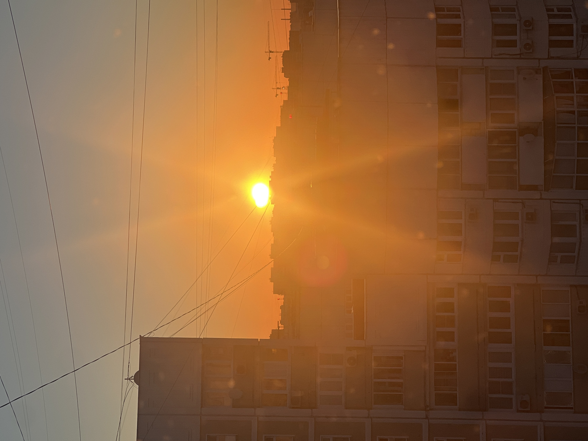 The image captures a city skyline during a bright sunset. The sun, a glowing orb of intense yellow and orange, is positioned centrally in the frame, casting a warm, radiant light across the scene. Sunlight streaks emanate from the sun, creating a lens flare effect that diffuses the light and adds to the ethereal quality of the sunset.
Dominating the left side of the image is a multi-story apartment building, its grey facade punctuated by rows of windows. The building appears to be of Soviet-era architecture, characterized by its functional design and panel construction. Above the building, thin black wires crisscross the sky, likely power lines.
In the distance, silhouetted against the vibrant sunset, are other buildings, forming a dense urban landscape. Several construction cranes are visible among these distant structures, suggesting ongoing development within the city. The sky transitions from a pale blue at the top to a deep orange and yellow near the horizon, a typical color gradient during a sunset. The overall impression is one of urban life bathed in the dramatic, fleeting beauty of the setting sun. While the image provides a sense of urban environment, specific details that would pinpoint a precise location within Moscow, Russia, such as unique architectural landmarks or street signs, are not discernable.