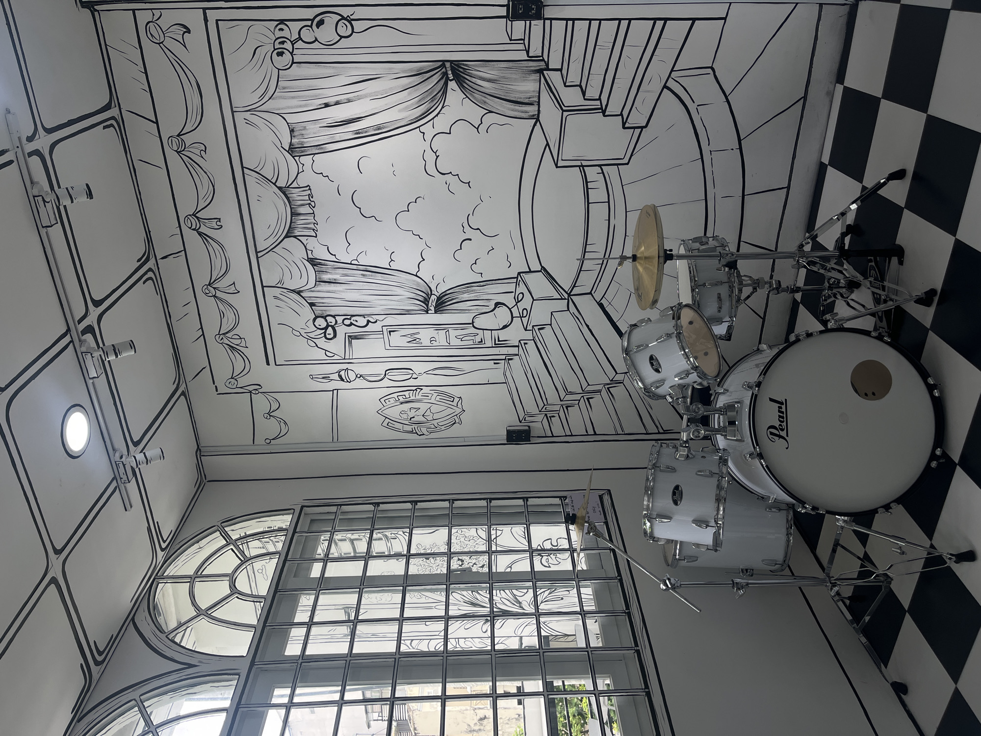 The image depicts the interior of a room in Ho Chi Minh City, Vietnam, designed to resemble a 2D comic book. The walls and ceiling are adorned with black line drawings that create the illusion of a cartoon environment. The artwork features a theatrical stage with curtains and clouds, a grand staircase, and architectural details like windows with intricate grilles. In the foreground, a white drum set, with cymbals and stands, is positioned on a black and white checkered floor. The room is illuminated by a ceiling light. The overall scene presents a unique, artistic ambiance.