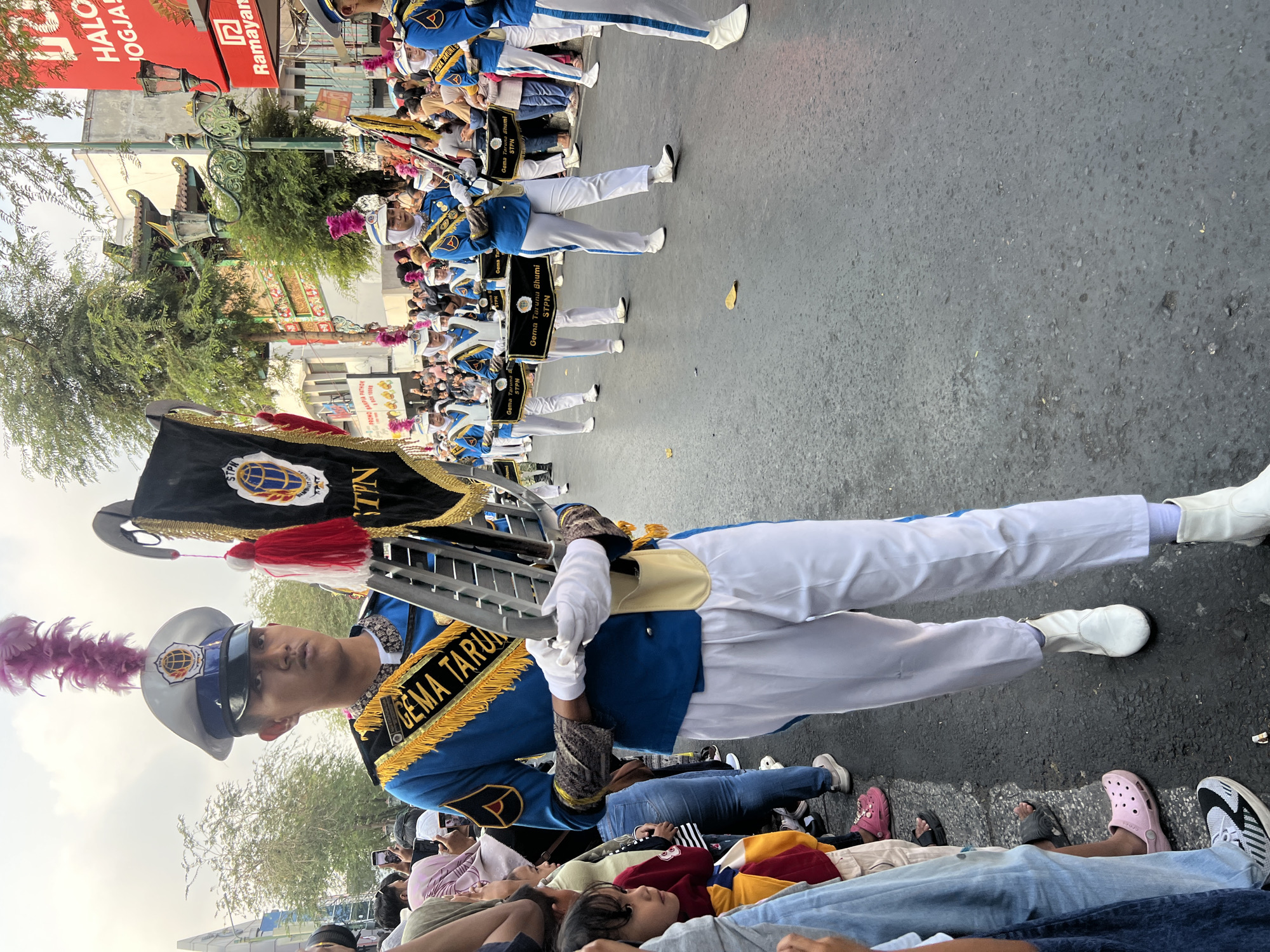 A parade is taking place on a street in Yogyakarta, Indonesia. The foreground features a young person in a marching band uniform, consisting of a blue jacket with gold trim and epaulets, white pants, and a white hat adorned with a purple feather. They are holding a banner with gold fringe and tassels, as well as a part of a marching instrument.
In the background, other members of the marching band are visible, also in uniform and carrying drums. Spectators line the street, some taking pictures with their phones. Buildings with traditional Javanese architectural elements and modern signage are visible along the street. The street is paved and appears wet in places. The sky is overcast, suggesting it might be daytime. The overall scene depicts a cultural event or celebration in the city.