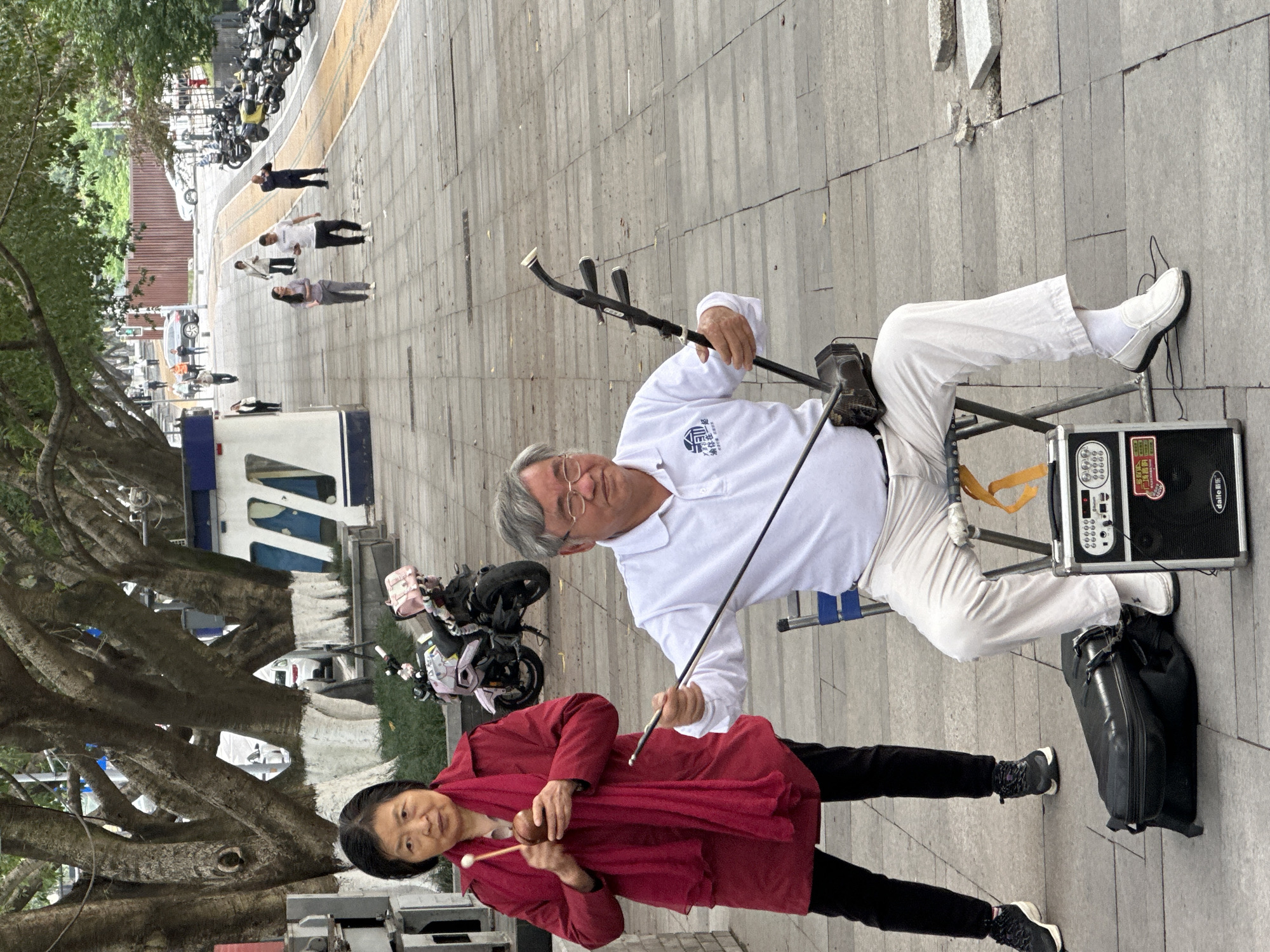 In Jiulongpo District, China, an elderly man sits and plays an Erhu, with an amplifier and black bag at his feet. He is wearing a white shirt and white pants, with white shoes and socks. A woman stands to his left, wearing a red coat, black pants, and black shoes. She is holding a pair of percussion instruments. Behind them is a large tree and a row of parked motorcycles. In the background, people walk along the sidewalk.