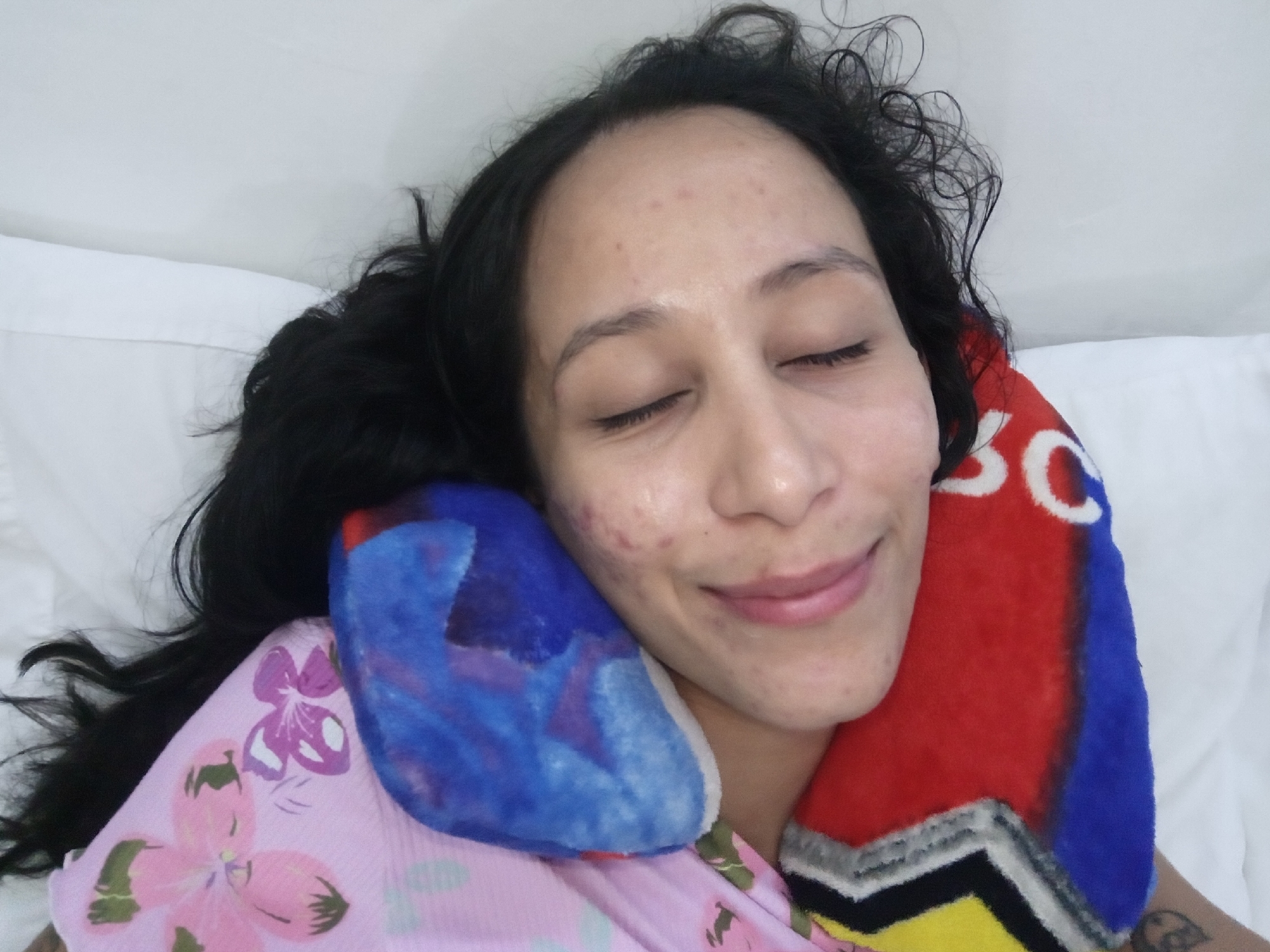A woman with dark hair is lying down with her eyes closed and a slight smile. She is on a white pillow or bed. She is wearing a pink floral top and is resting her head on a colorful neck pillow with red, blue, and yellow sections. Part of a tattoo is visible on her right arm. The image does not provide information about the city of Porlamar, Venezuela.