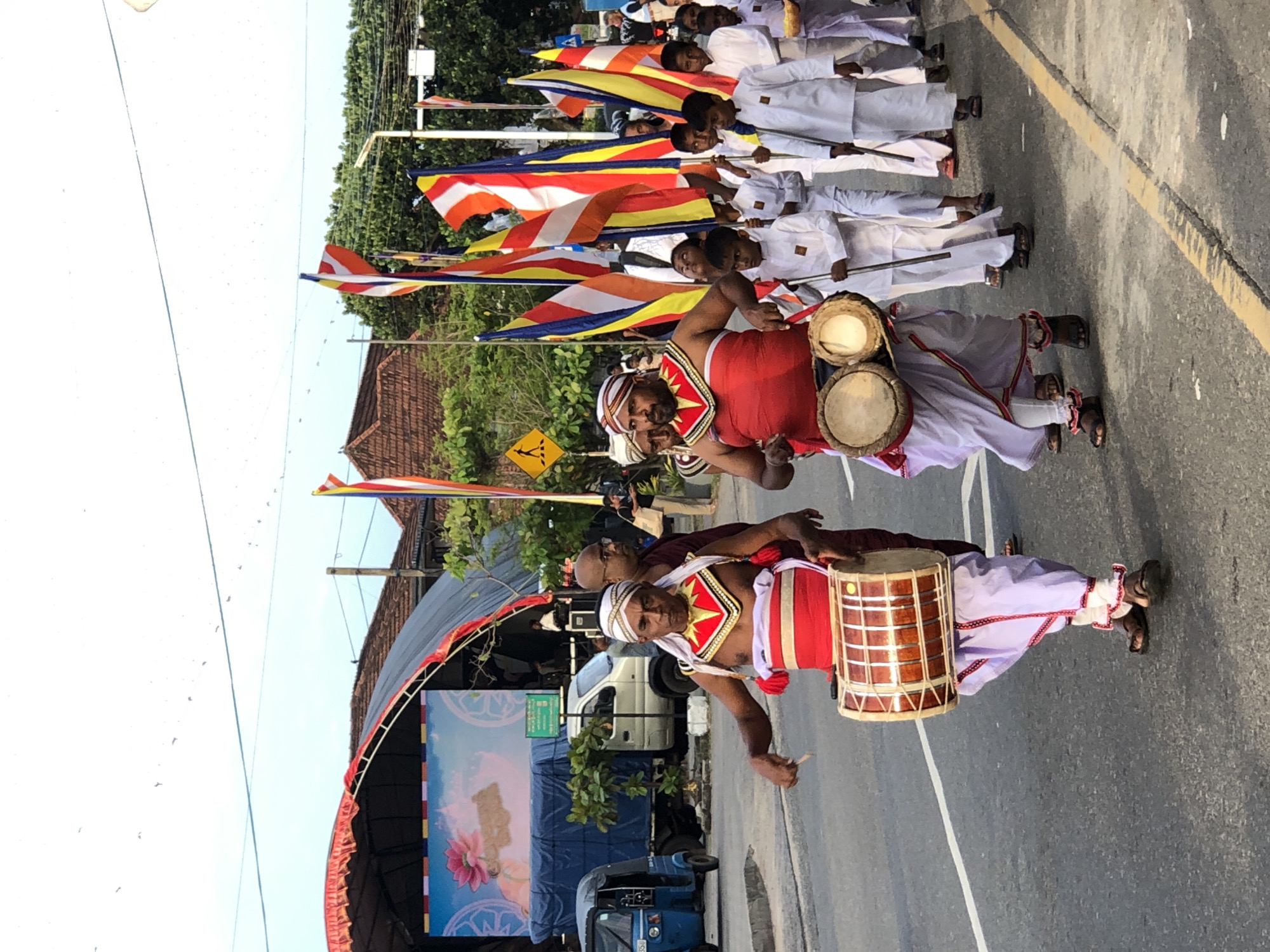 In Habaraduwa, Sri Lanka, a procession moves along a paved road under a bright sky with scattered birds. In the foreground, two men, dressed in traditional white and red ceremonial attire with elaborate headbands and chest adornments, are playing drums. The man on the left holds a large, cylindrical drum horizontally, while the man on the right plays a set of two smaller, hourglass-shaped drums suspended from his waist. Both men are actively engaged in drumming, with sticks in hand.
Behind them, a group of boys, also in white attire, follow carrying long sticks, possibly part of a ceremonial guard or participants. In the background, several Buddhist flags, featuring stripes of orange, yellow, red, and blue, are visible, adding to the festive or religious atmosphere. Beyond the procession, a stage structure with a large screen displaying an image of a lotus flower is set up, with a blue truck and a small three-wheeled vehicle parked nearby. Buildings with tiled roofs and trees line the side of the road, indicating an urban or semi-urban setting. The overall scene suggests a cultural or religious event taking place in the town.